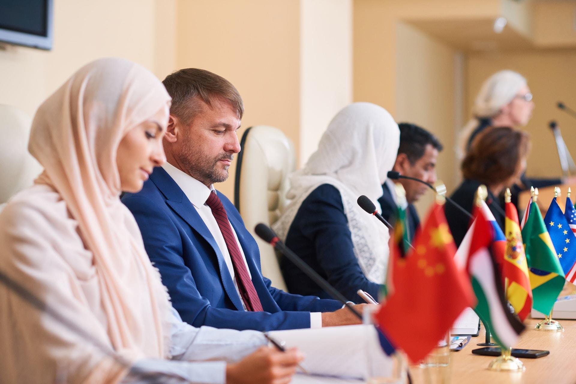 Group of people sitting in a brightly-coloured room at a long table, each with a flag representing their nation in front of them.