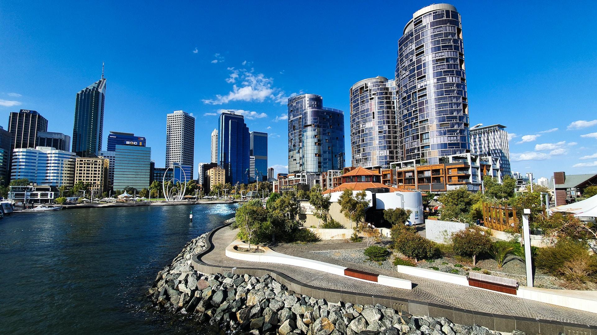 A skyline view of Perth featuring silver highrise buildings contrasting with a deep blue sky, with a body of water in the foreground.