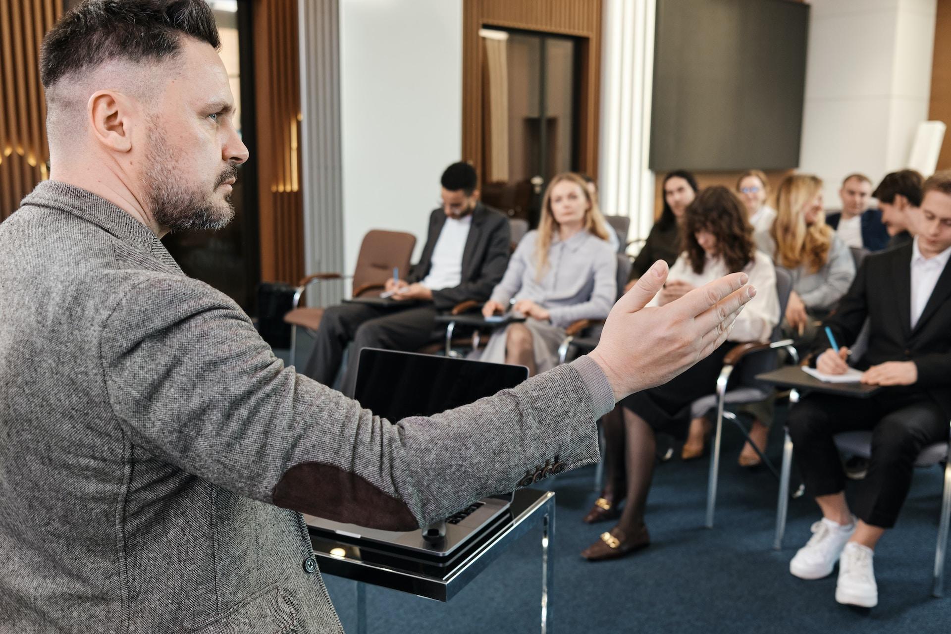A person in a grey tweed jacket with elbow patches stands next to a podium, before a group of people sitting in loosely arranged chairs with writing tools held in their laps.