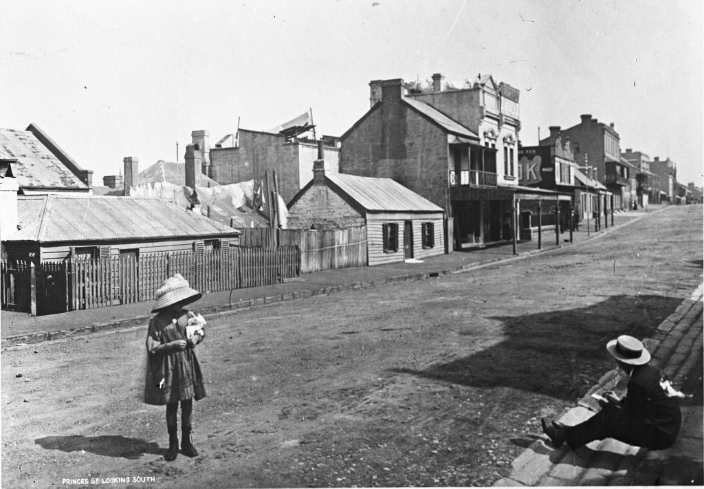 A black and white photo of a dirt street lined with single-storey buildings, and a small girl standing apart from an older boy, who is sitting down on the sidewalk