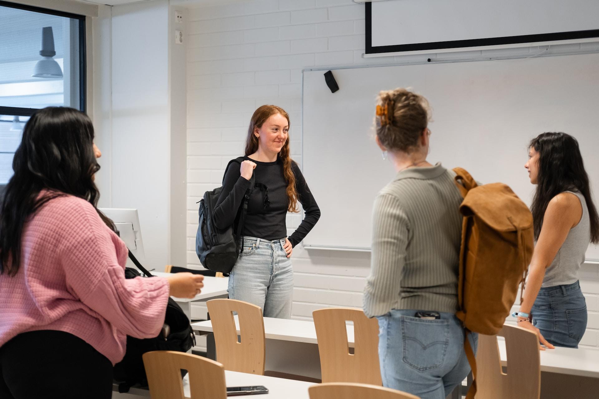 Four students stand in a classroom that has white table tops and wooden chairs, with backpacks slung over one shoulder, apparently talking about something. 