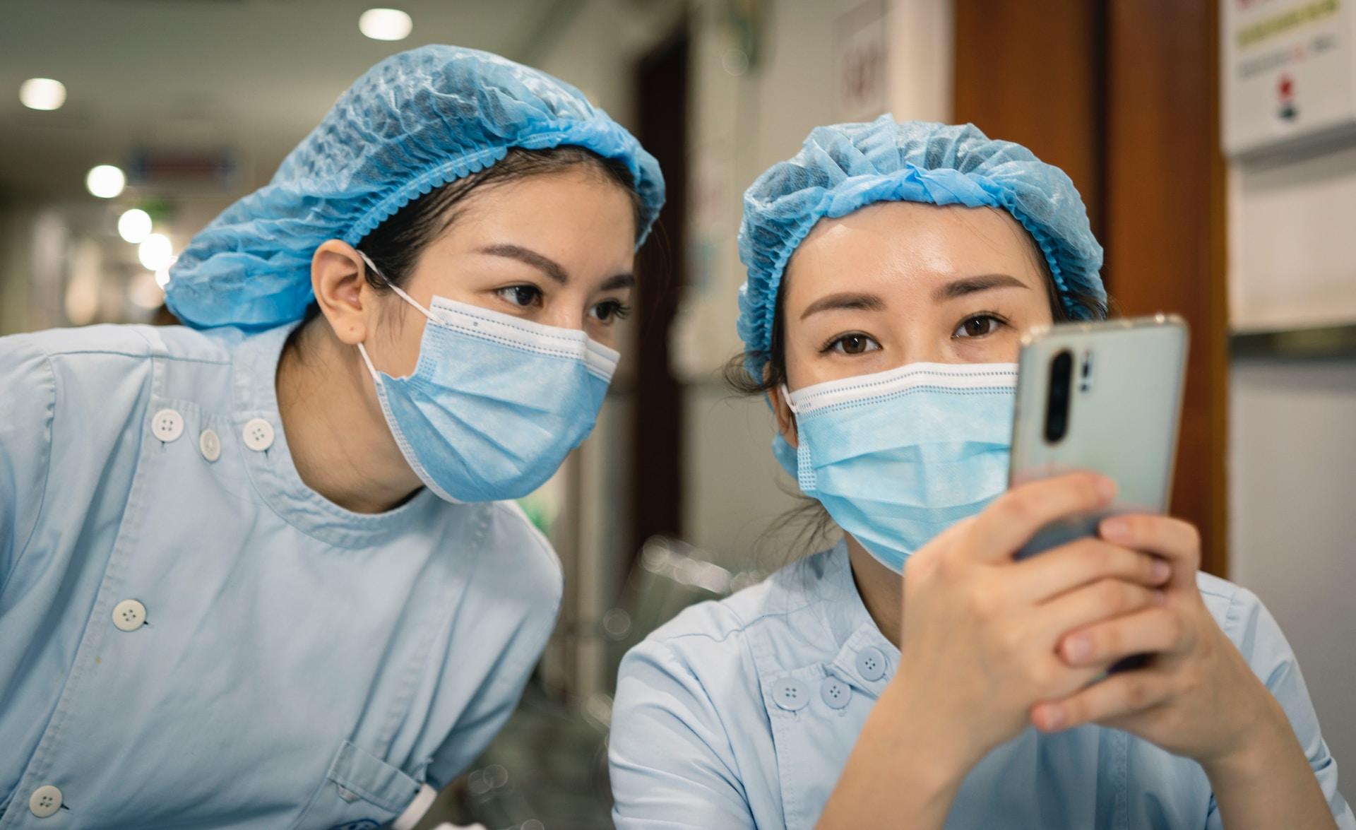 Two people wearing light green surgical scrubs, blue face masks and hairnets, stand close together, with one holding a smartphone and the other looking at its screen.