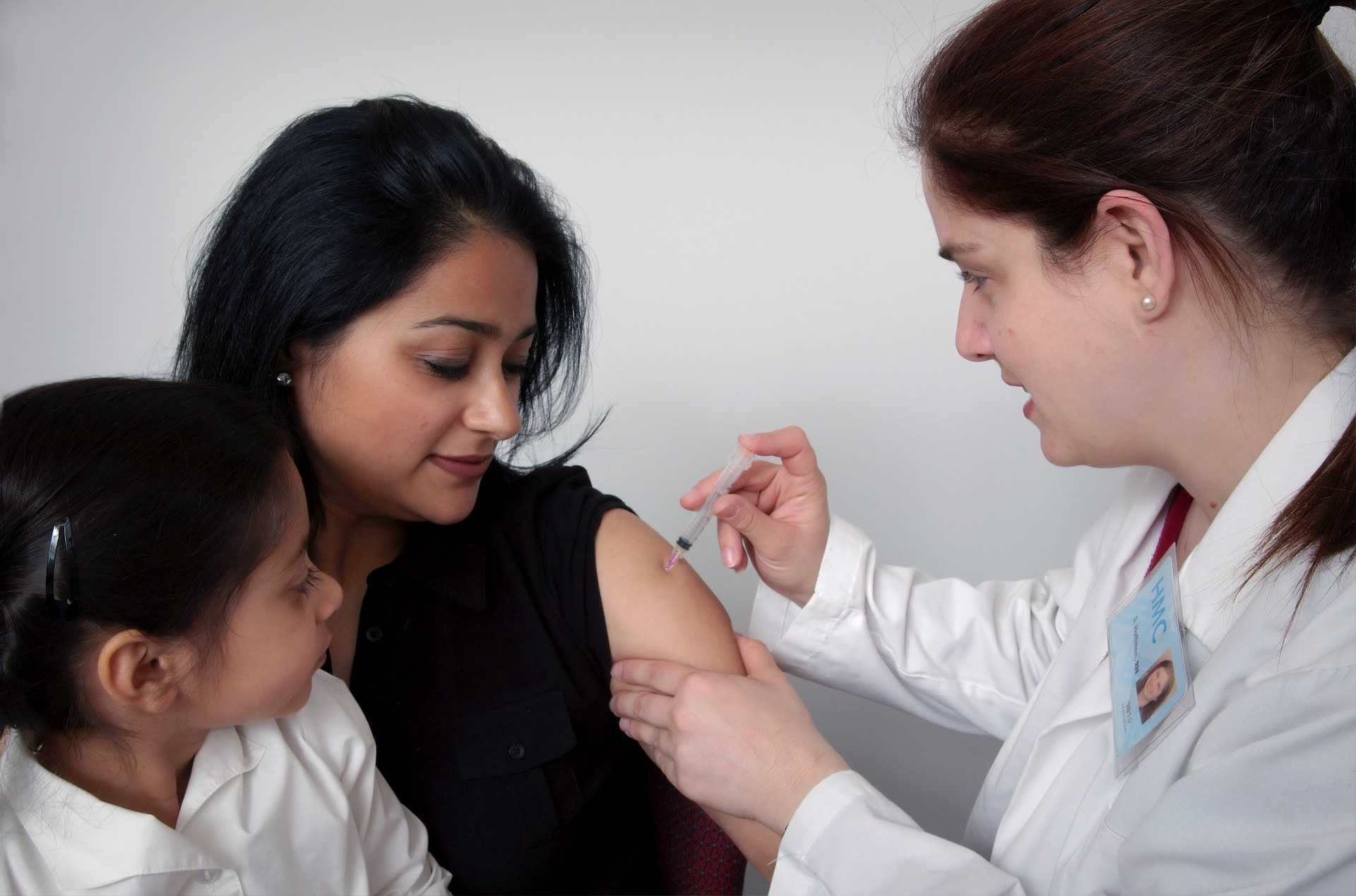 A person in a white lab coat administers an injection to a patient who has a young child sitting on their lap.