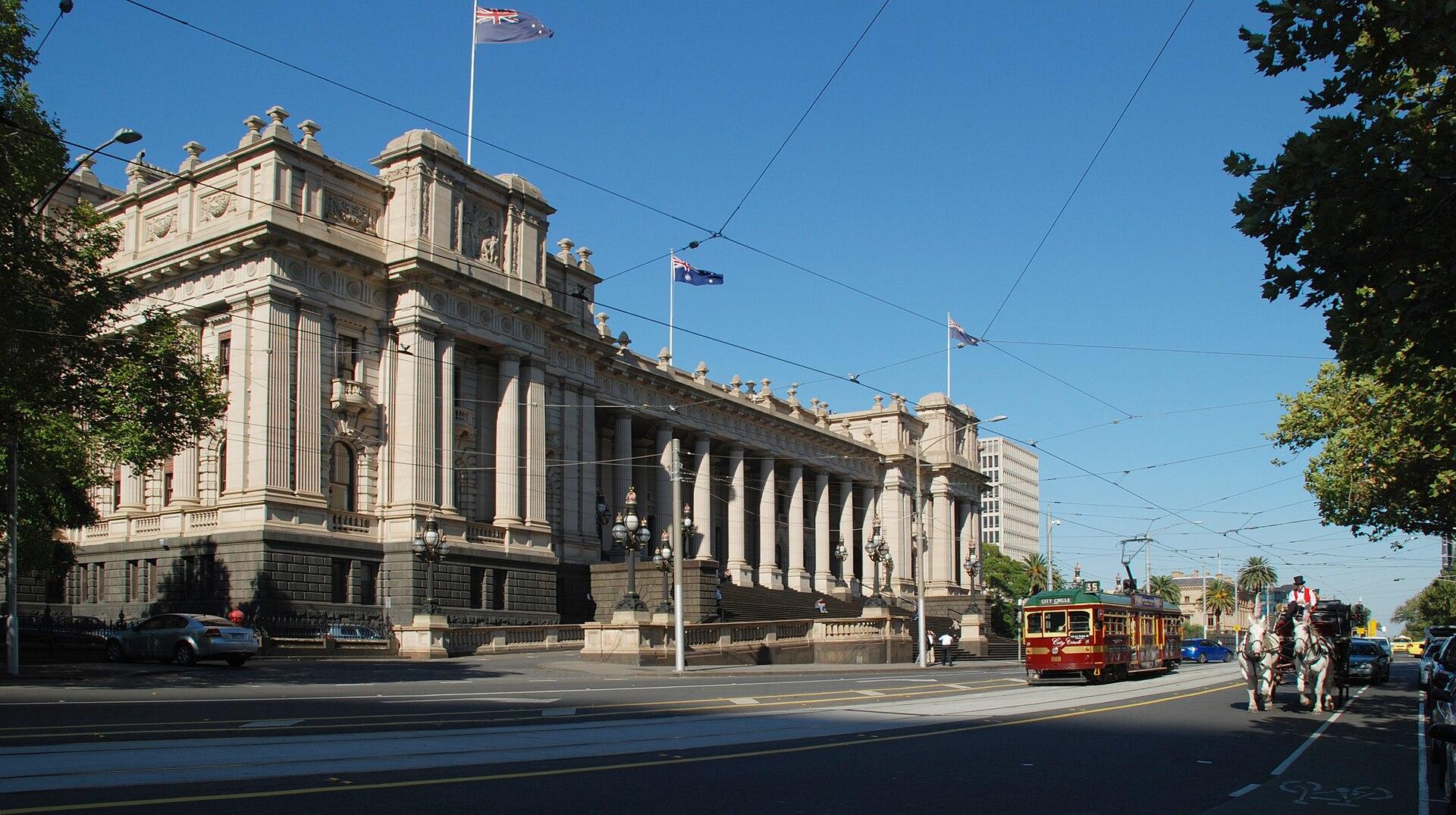 Victorian Parliament House, a large sandstone building done in Victorian style, with flags flying from the pinnacles and a blue sky above.