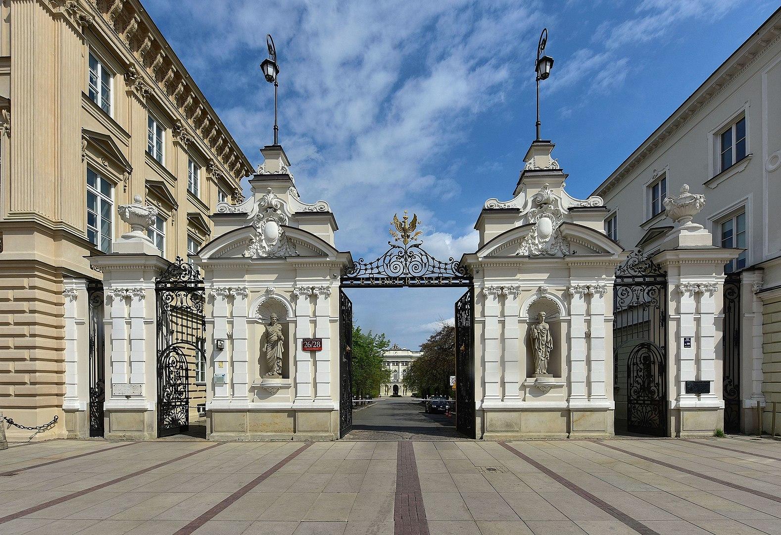 The main entry gates to Warsaw University appear white in the sunshine, while venerable sandstone buildings flank it on either side and a pavestone avenue entices strollers.