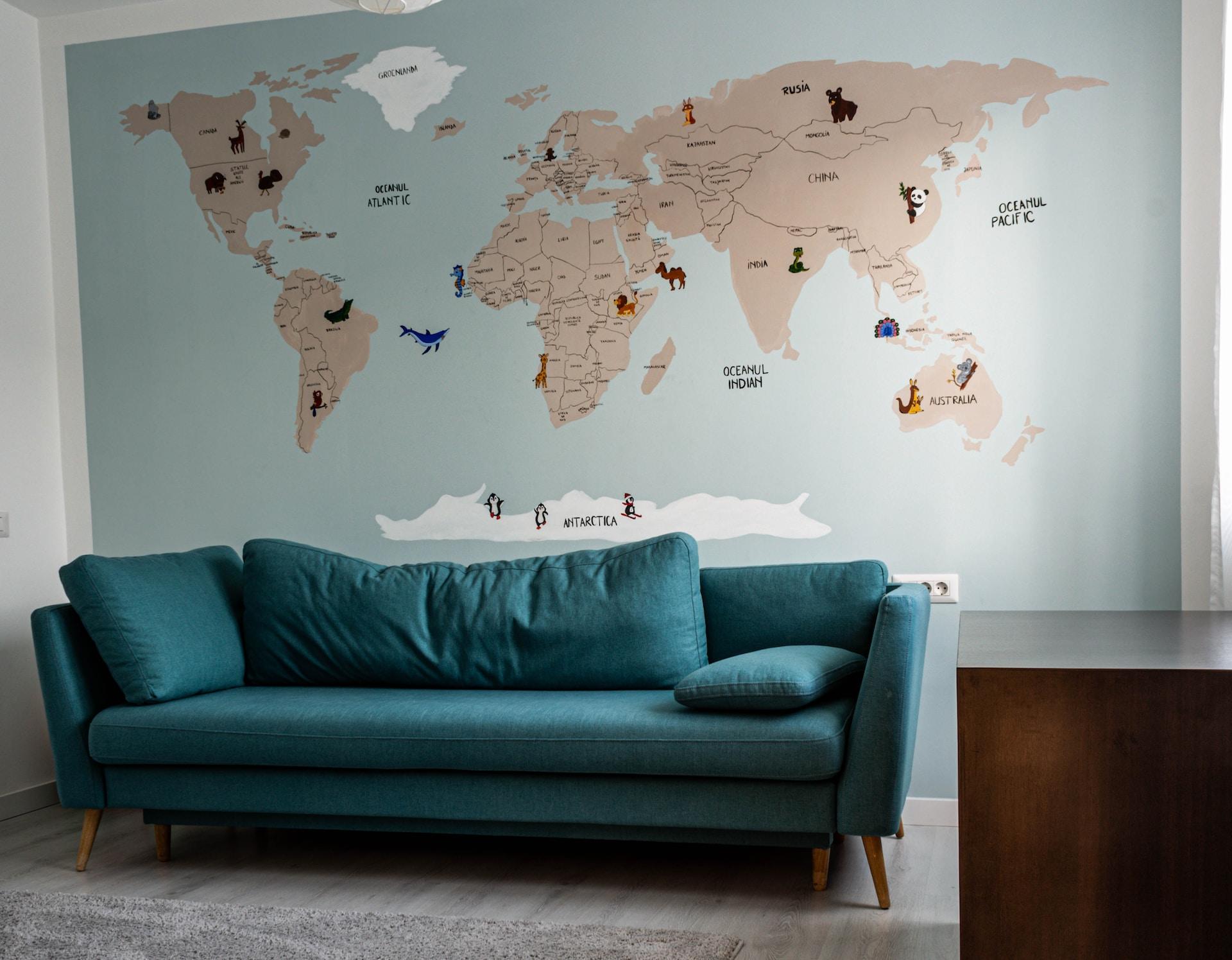 A mural depicting a world map with all the landmasses in brown and the arctic regions in white, set against a light blue background, with a turquoise coloured sofa in front of it.