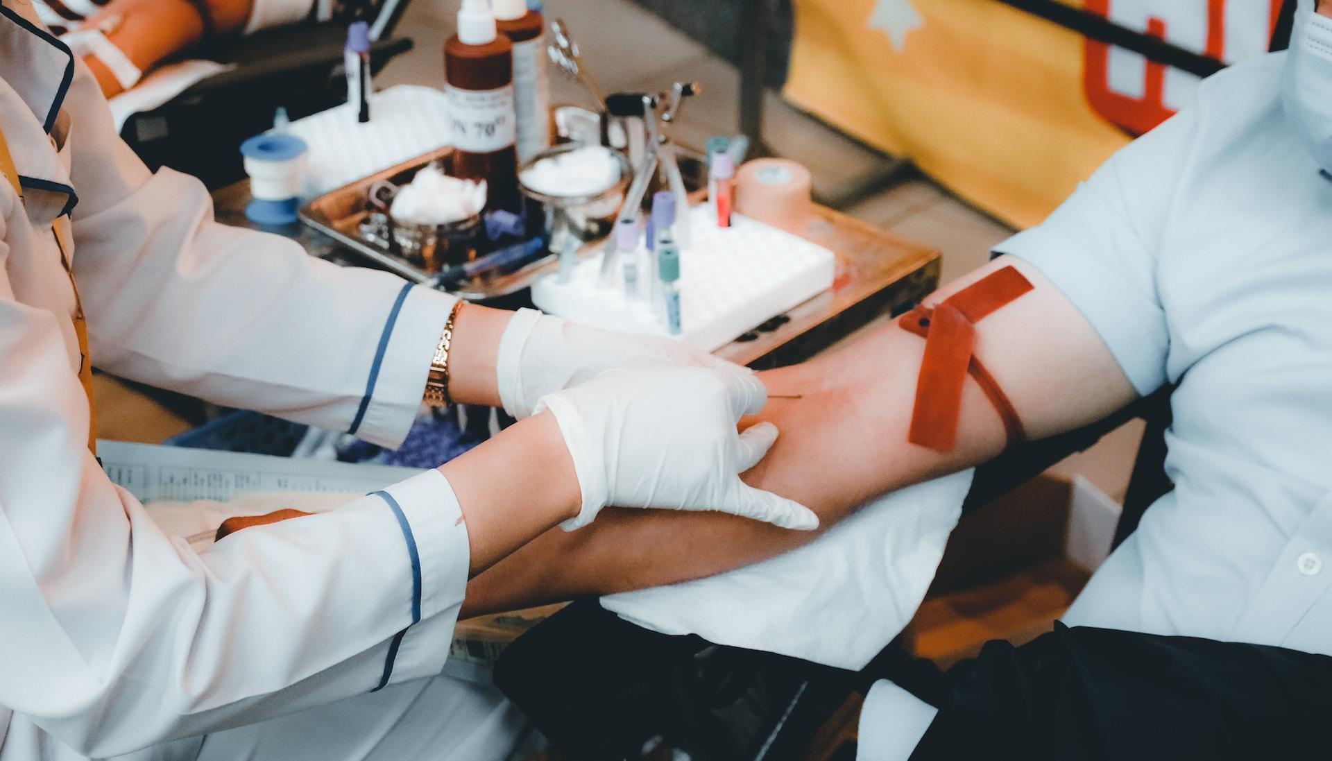 A person wearing white medical gloves prepares to draw blood from a patient's right arm, which is tied off with a red latex strip.