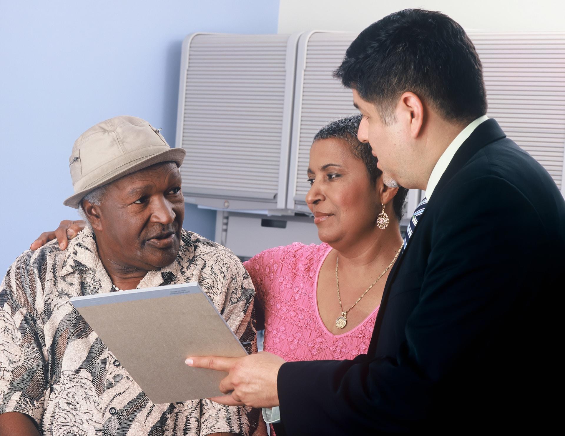 A person in a black business suit holds a legal pad up so two people sitting close to each other can see it. The person in the pink top has their arm around the person wearing a brown hat and patterned shirt. 