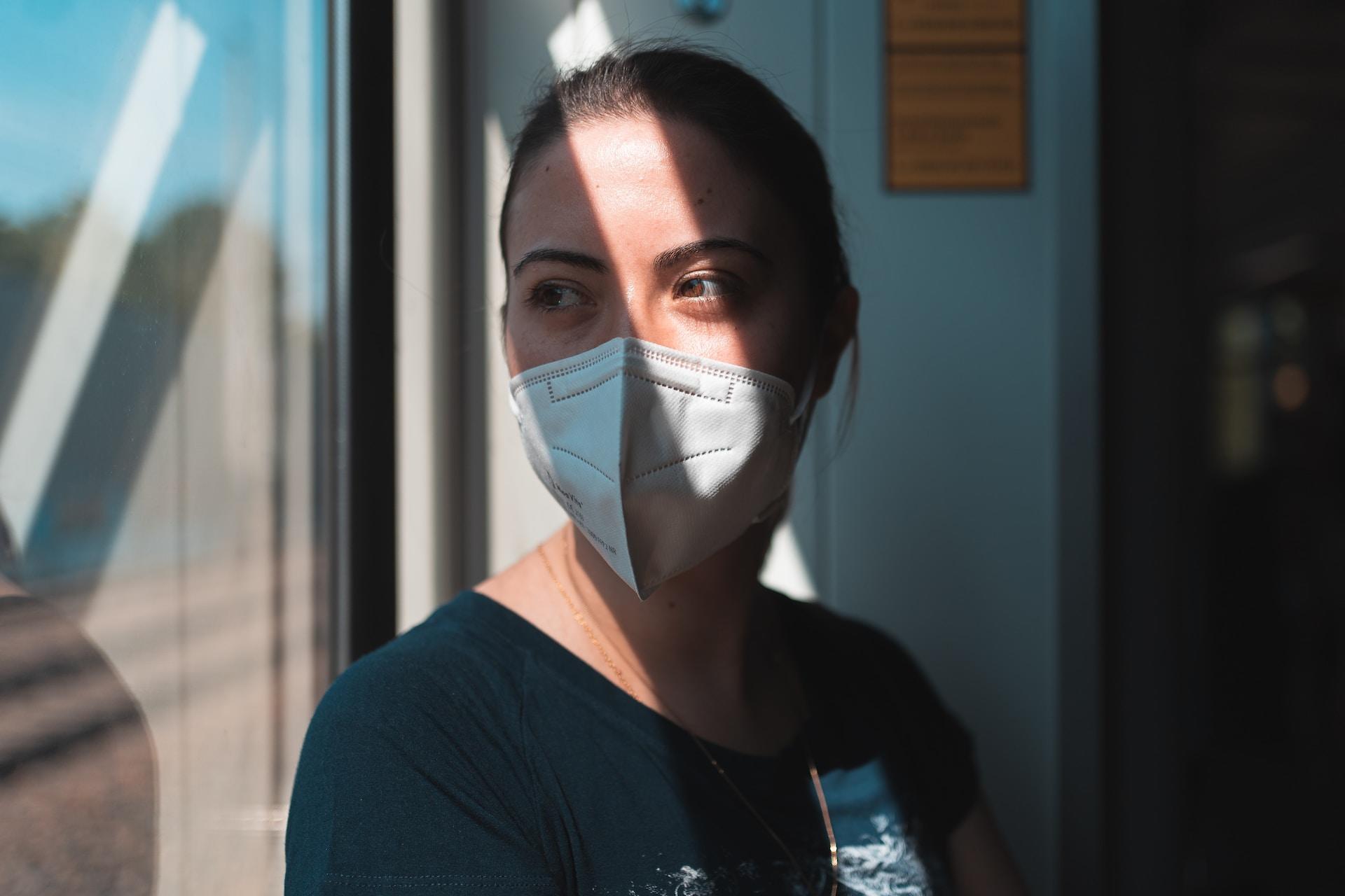 A person wearing a blue top and a white face masks stands by a window, letting the sun slash across their face. They look sad and pensive.