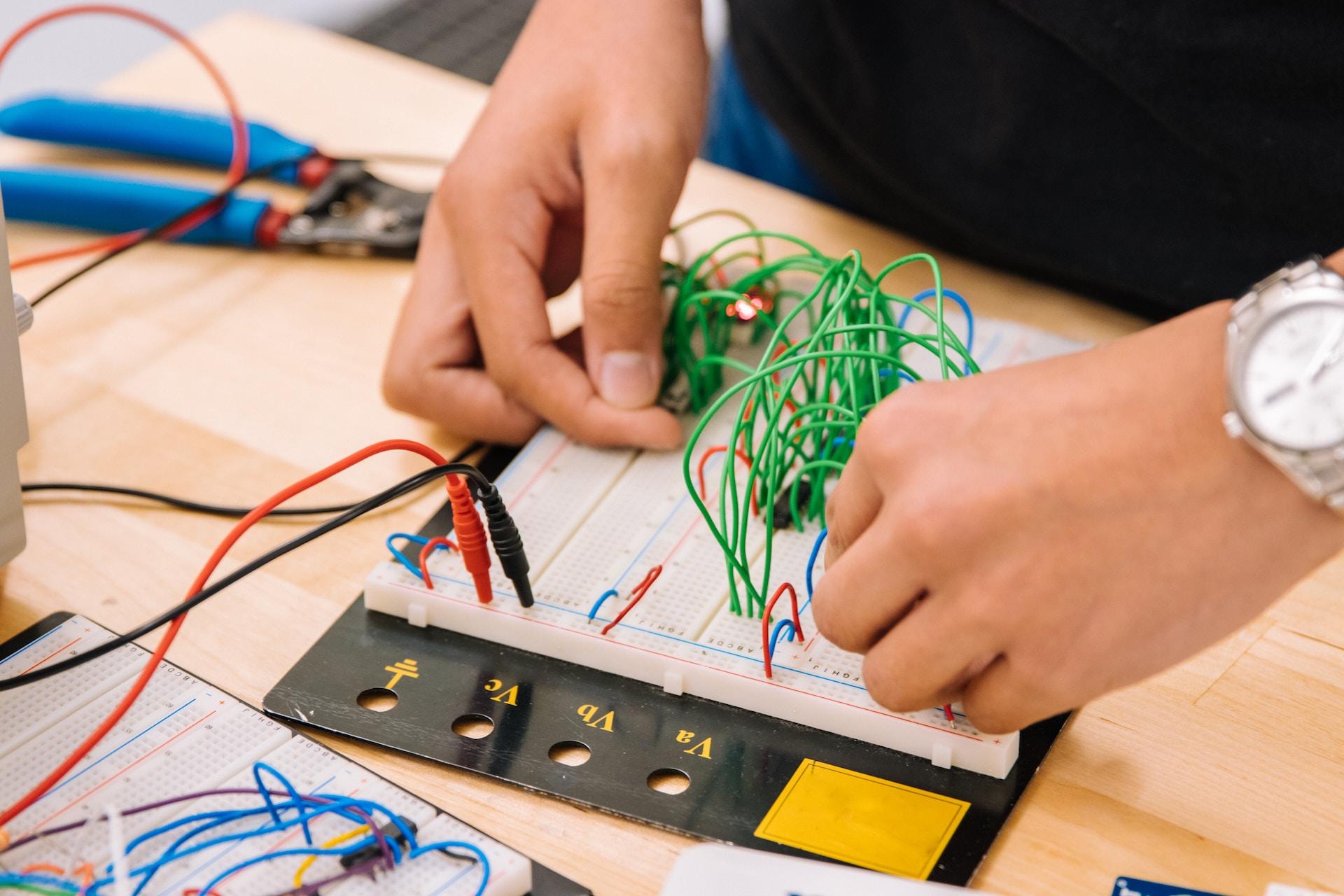 A person wearing a silver wristwatch stands at a workbend affixing green wires to a white circuit board. 