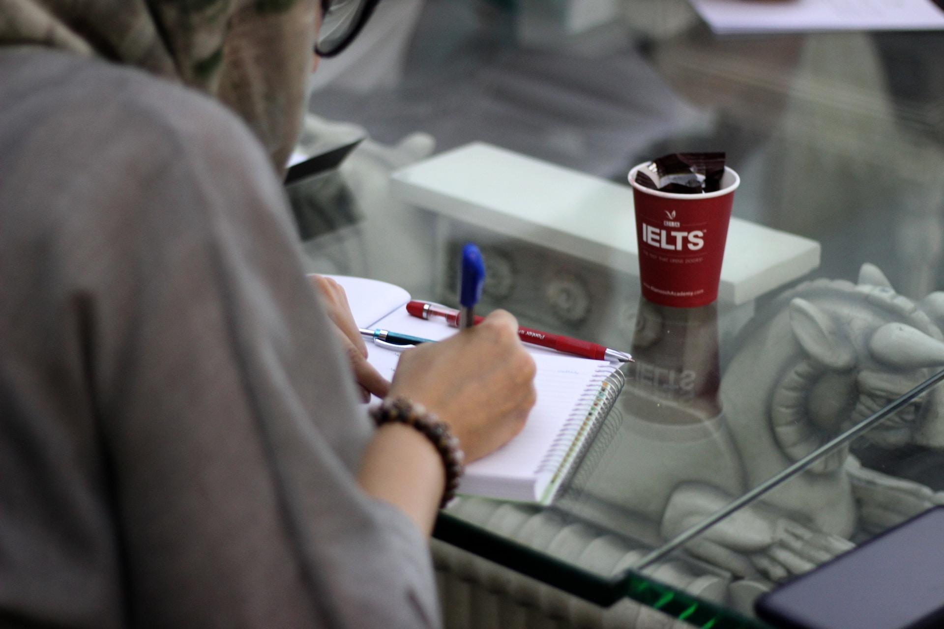 A disposable red hot-beverage cup with IELTS printed in beige sits on a glass table in front a a person's open notebook, wherein they're writing something by hand.
