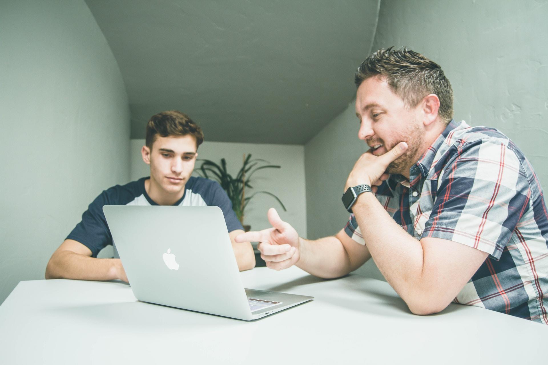 An older person in a short-sleeved checked shirt sits at a white-topped table, pointing at something on the open laptop computer's screen while a younger person wearing a blue and white tee shirt looks on.