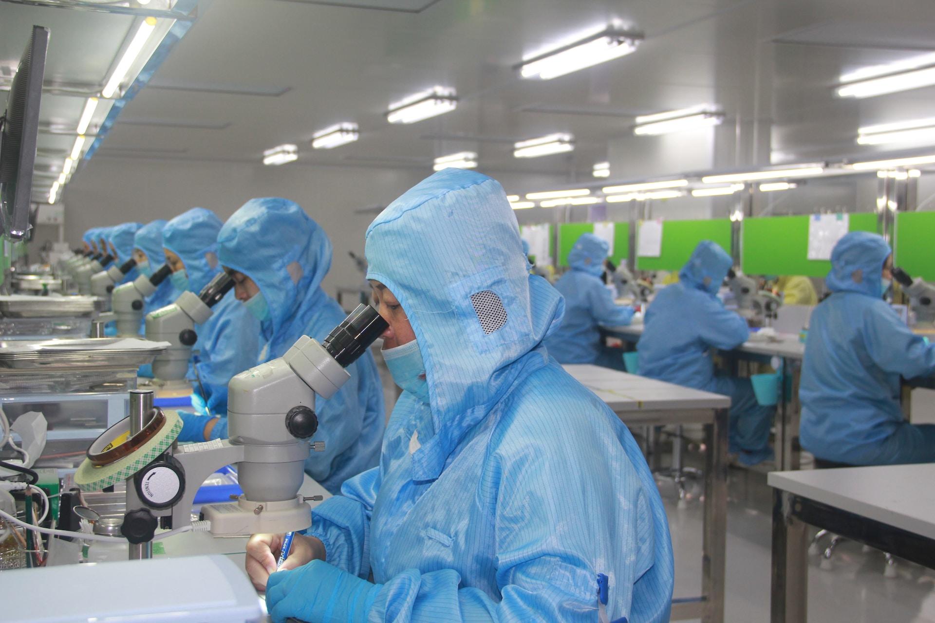 A long, brightly lit room with two rows of technicians wearing light blue protective suits with hoods and face masks, each sitting in front of a microscope, peer into the sights.