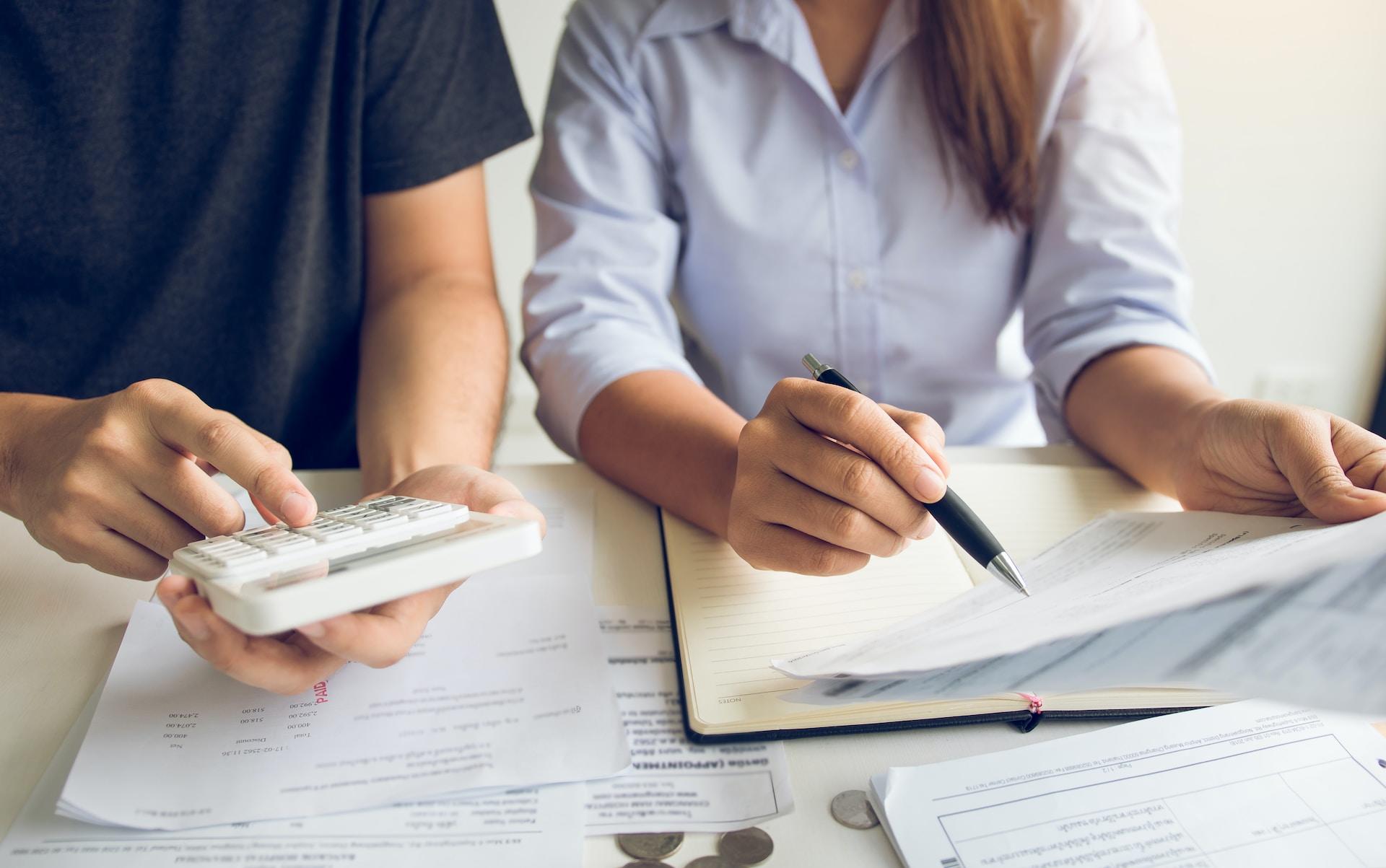 A person in a light-coloured blouse tilts a paper towards a person wearing a dark short-sleeved shirt who is holding a white calculator in their right hand. 