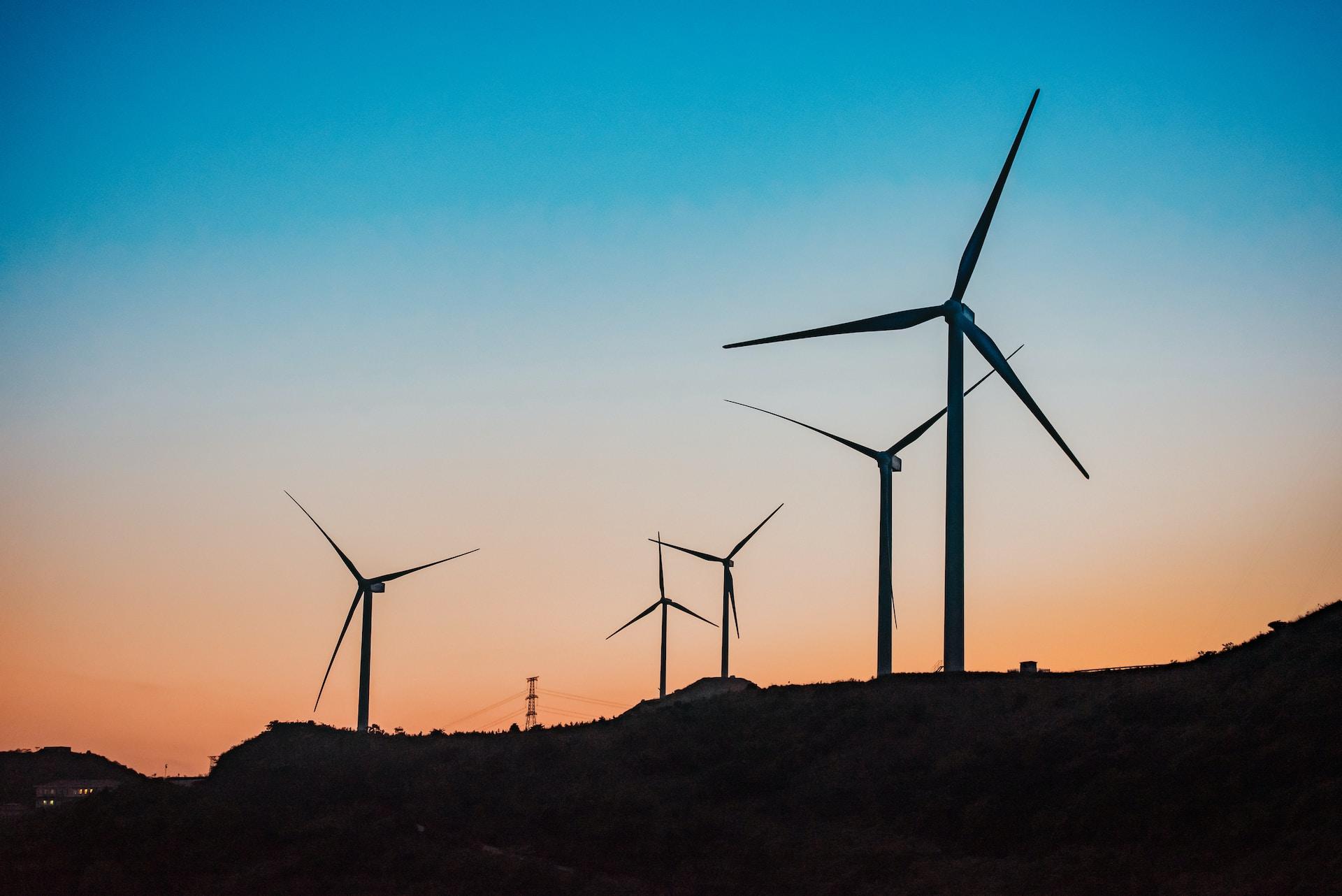 A windfarm on a hillside seen in silhouette against a colourful sunset sky with the hill already shrouded in darkness.