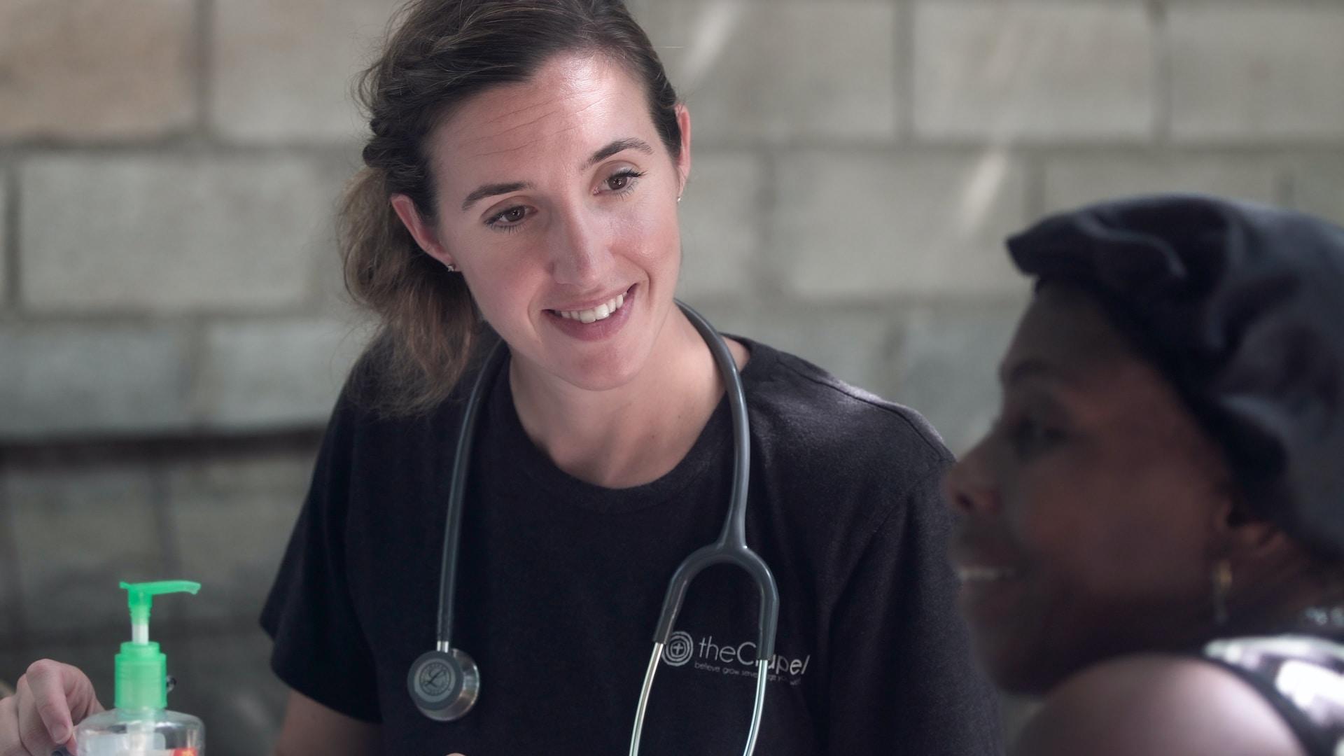 A person wearing a black top, with a stethoscope around their neck, smiles as they engage with a patient. 