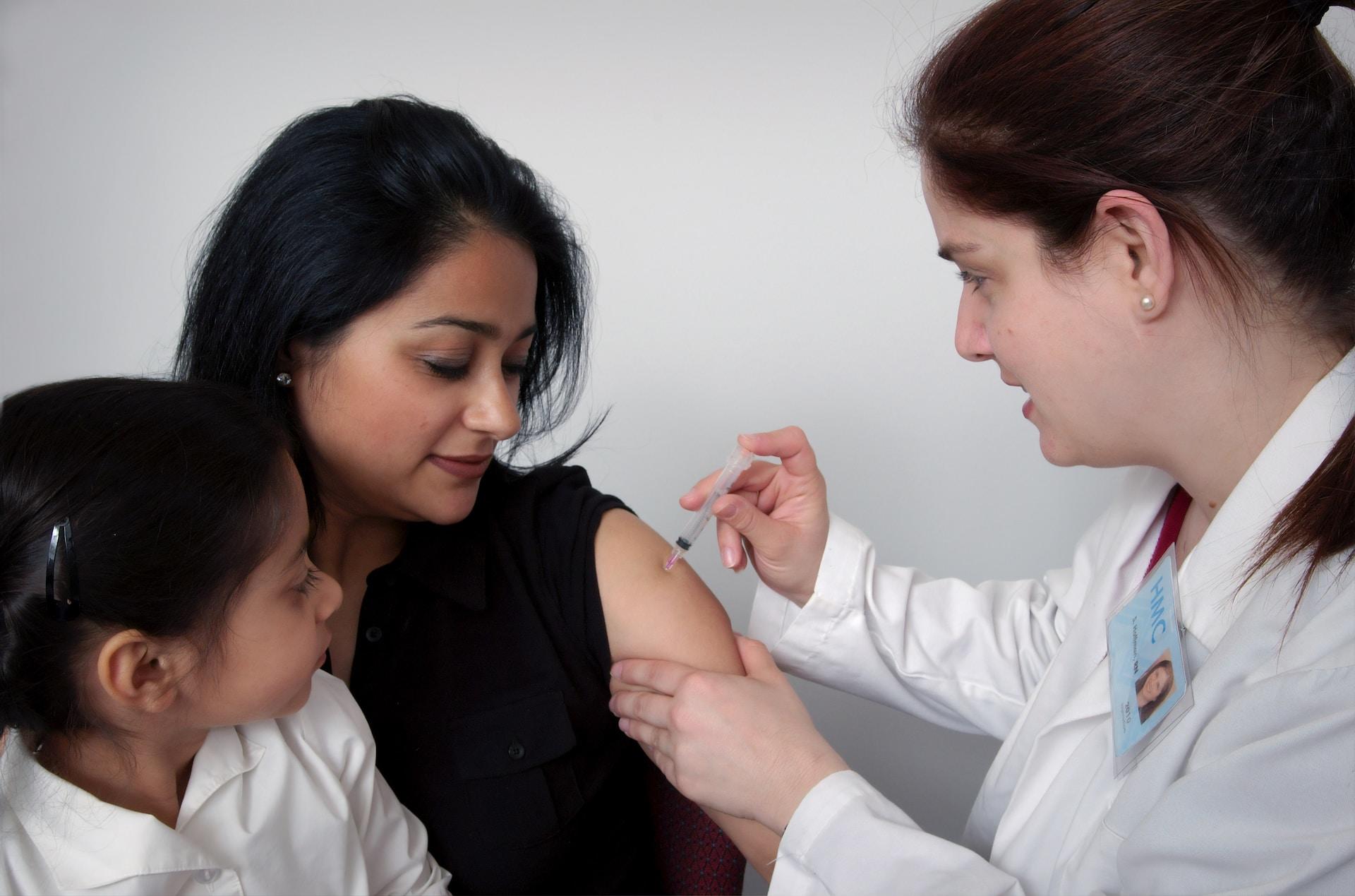 A person in a white lab coat administers an injection to a person wearing a black top with their sleeve rolled up while a young child wearing a white top sits on the patient's lap. 