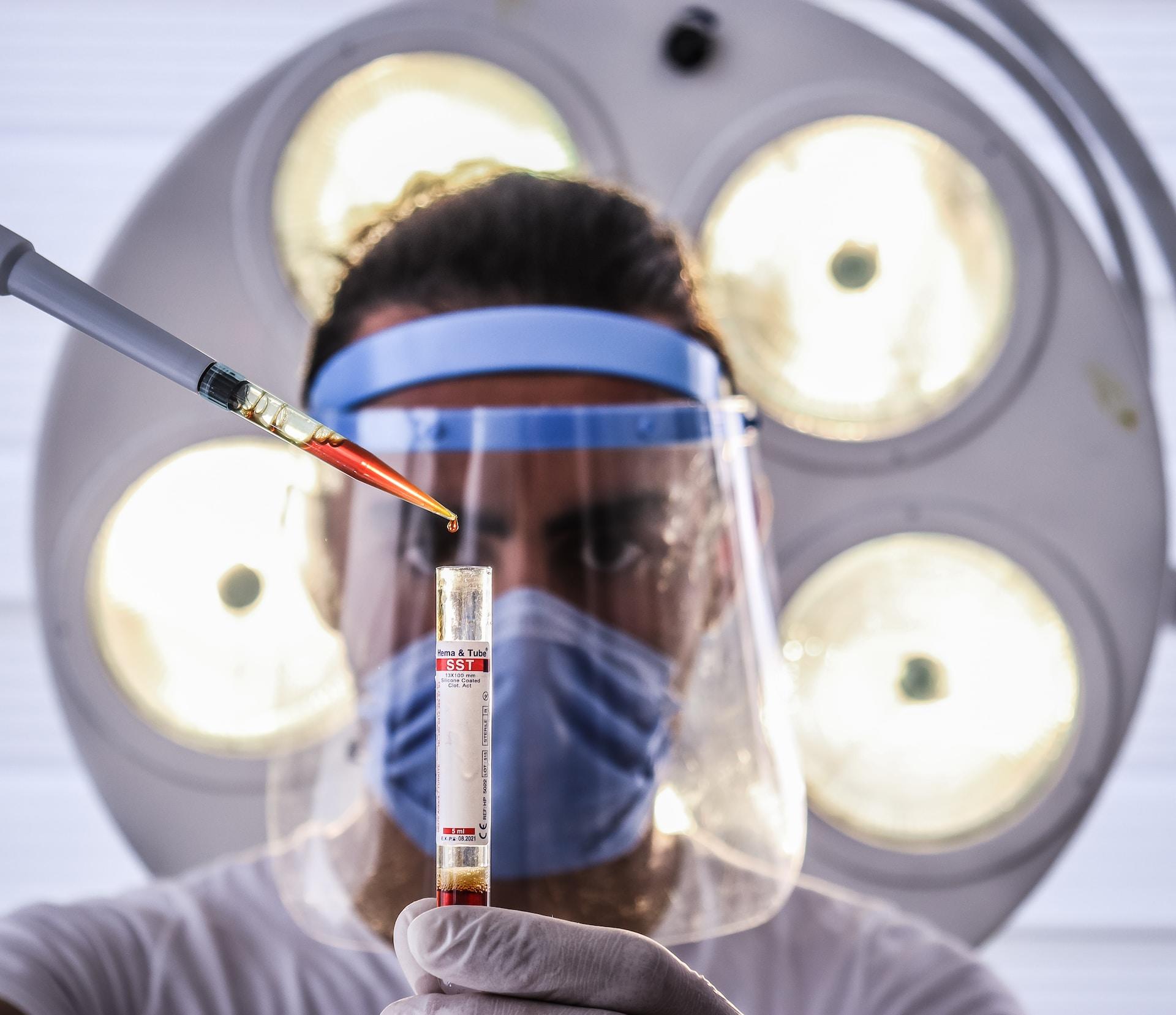 A person wearing a clear face shield with a blue headband and a medical mask underneath focuses on dripping a red liquid from a pipette into a test tube. 