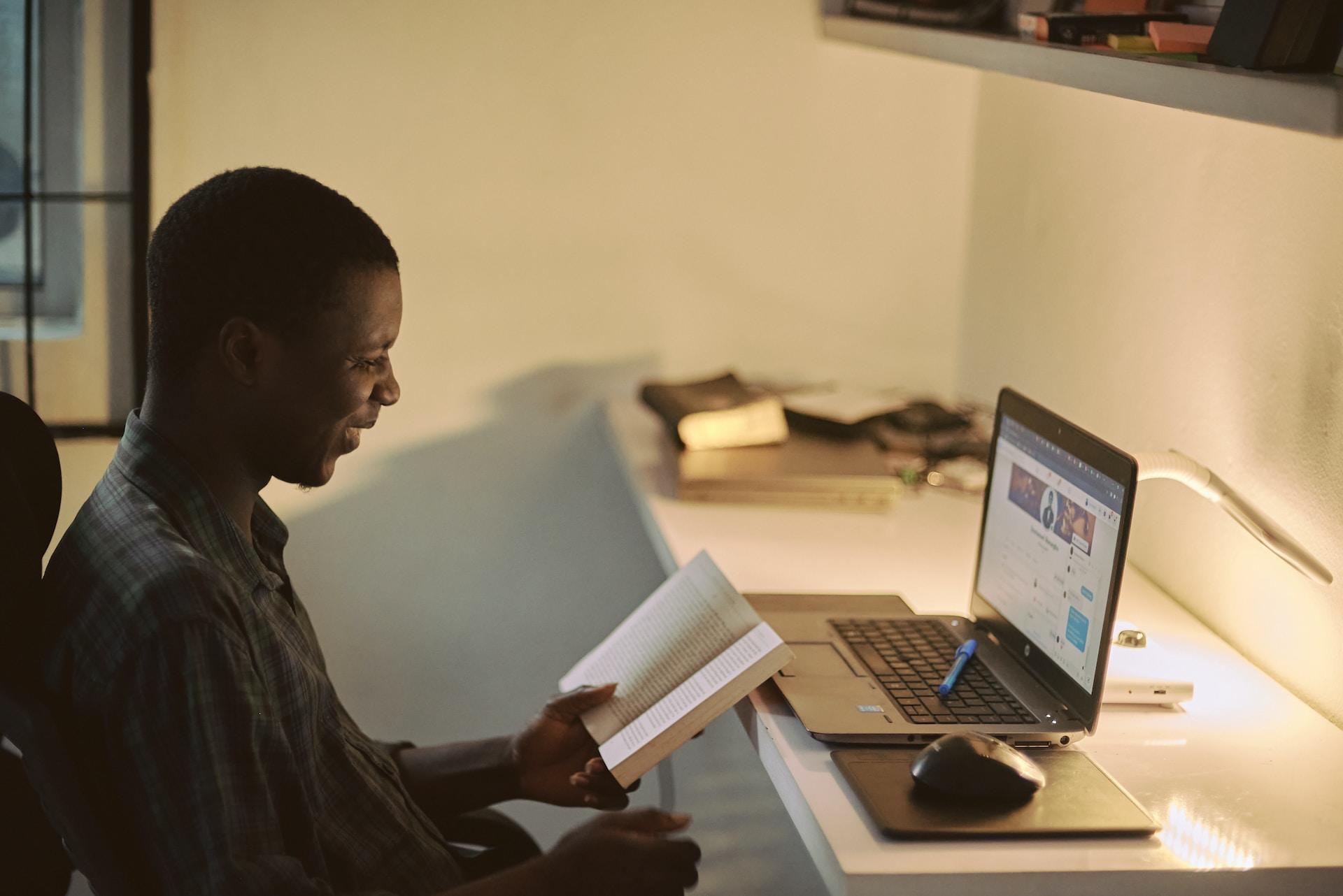 A person sits at a lit-up workspace in front of an open laptop computer with an open book in their left hand. 