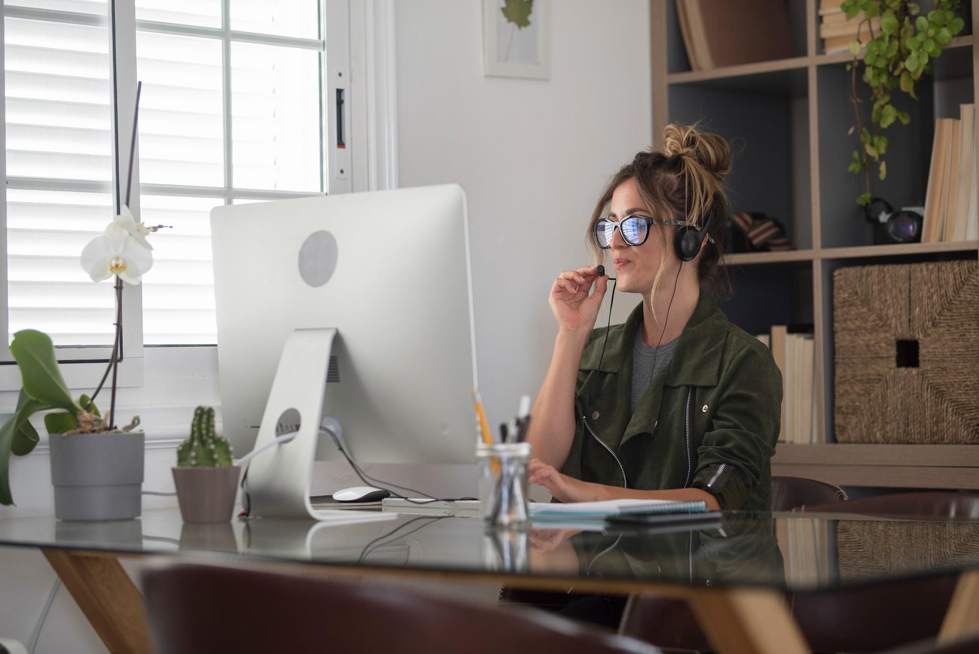 A person wearing a kakhi top and glasses sits at a glass desk in a well-appointed office, speaking into a boom microphone mounted on a headset during a videoconference.