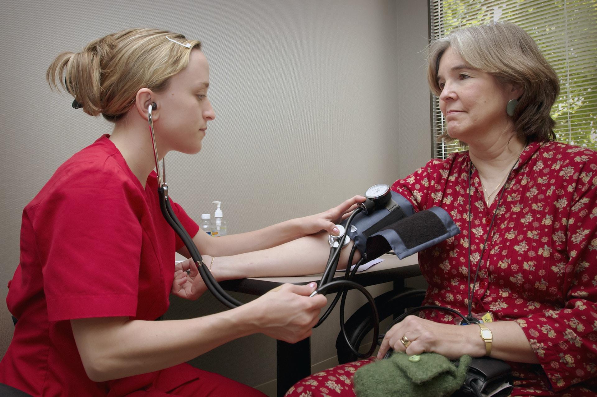 A person wearing bright red surgical scrubs monitors a blood pressure cuff on a patient's arm as the patient, wearing a dark red top with light coloured flowers stares vacantly ahead.