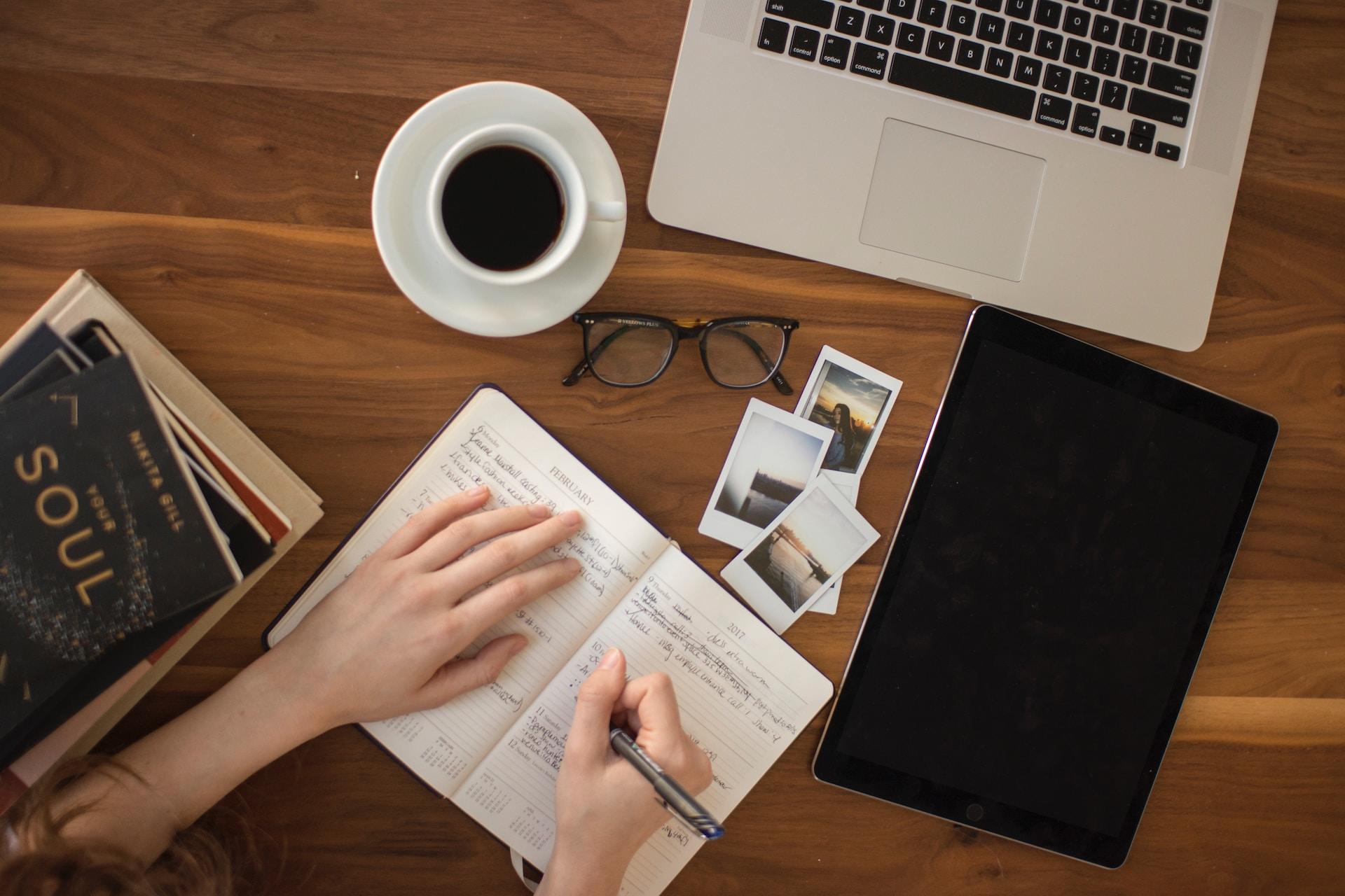 A person holding a pen in their right hand, poised over an open notebook. Their left hand rests on the left page and they have a pair of black-framed eyeglasses, a cup of coffee, some polaroid snaps and a laptop computer on the wooden table in front of them.