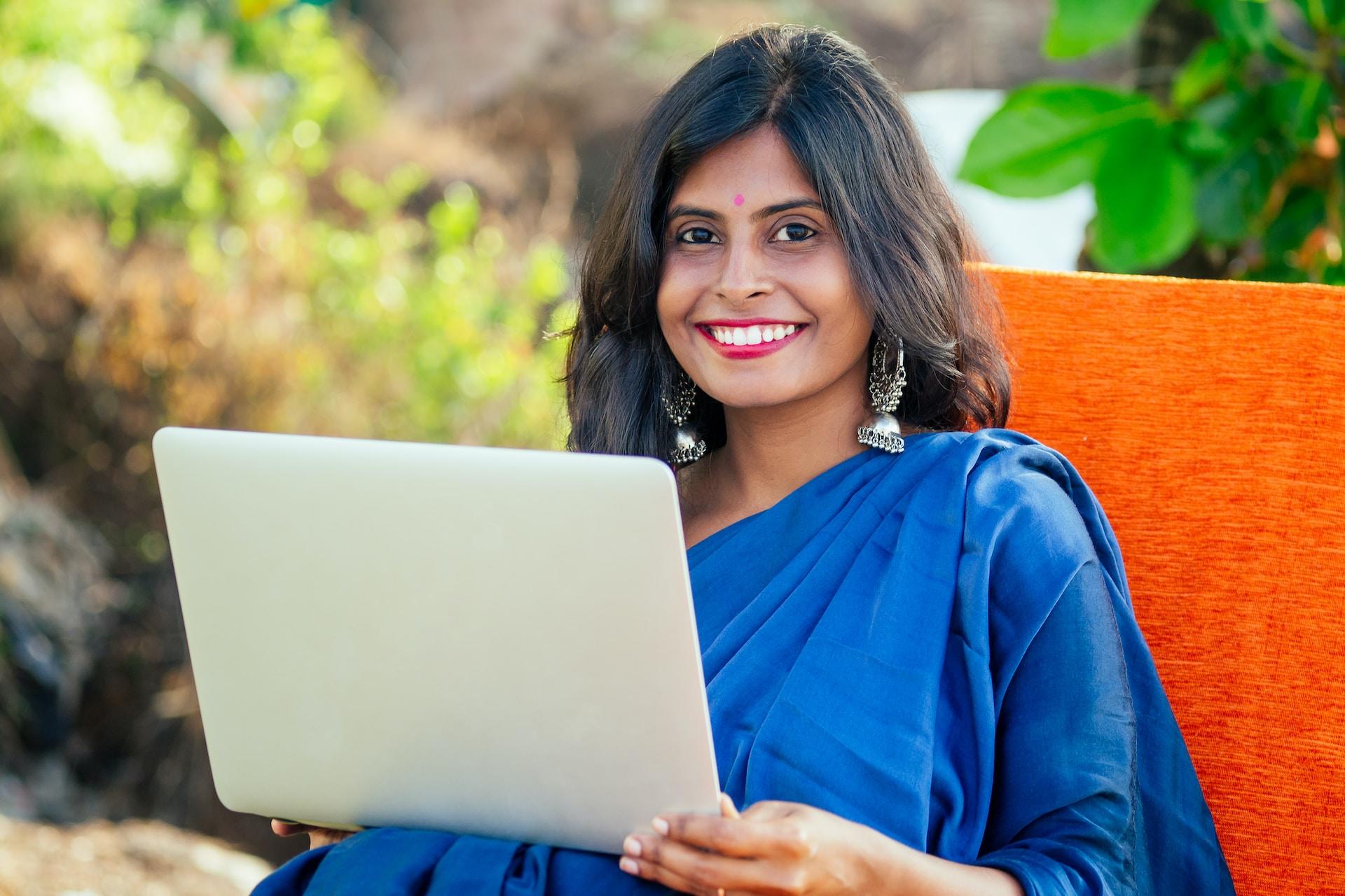A woman in a royal blue sari sits on an orange chair with a white tablet in her hands, smiling at the camera.
