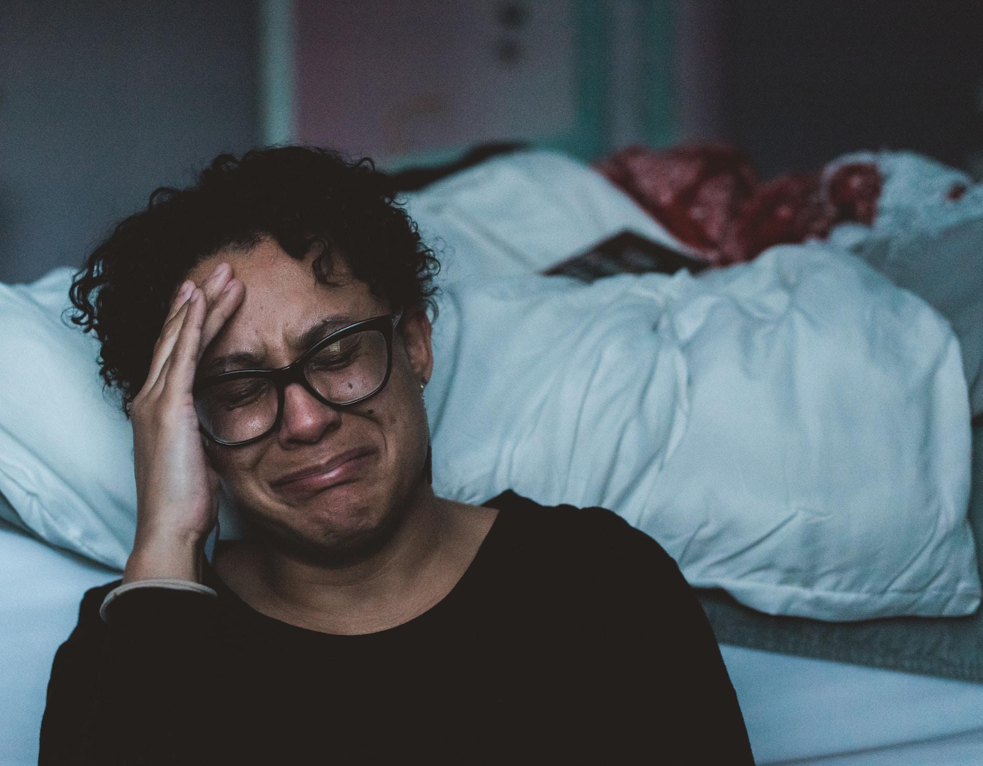 A woman wearing dark framed glasses and a dark coloured top sits on the floor and leans against an unmade bed with white linens, with her head in her hand and appearing to cry.