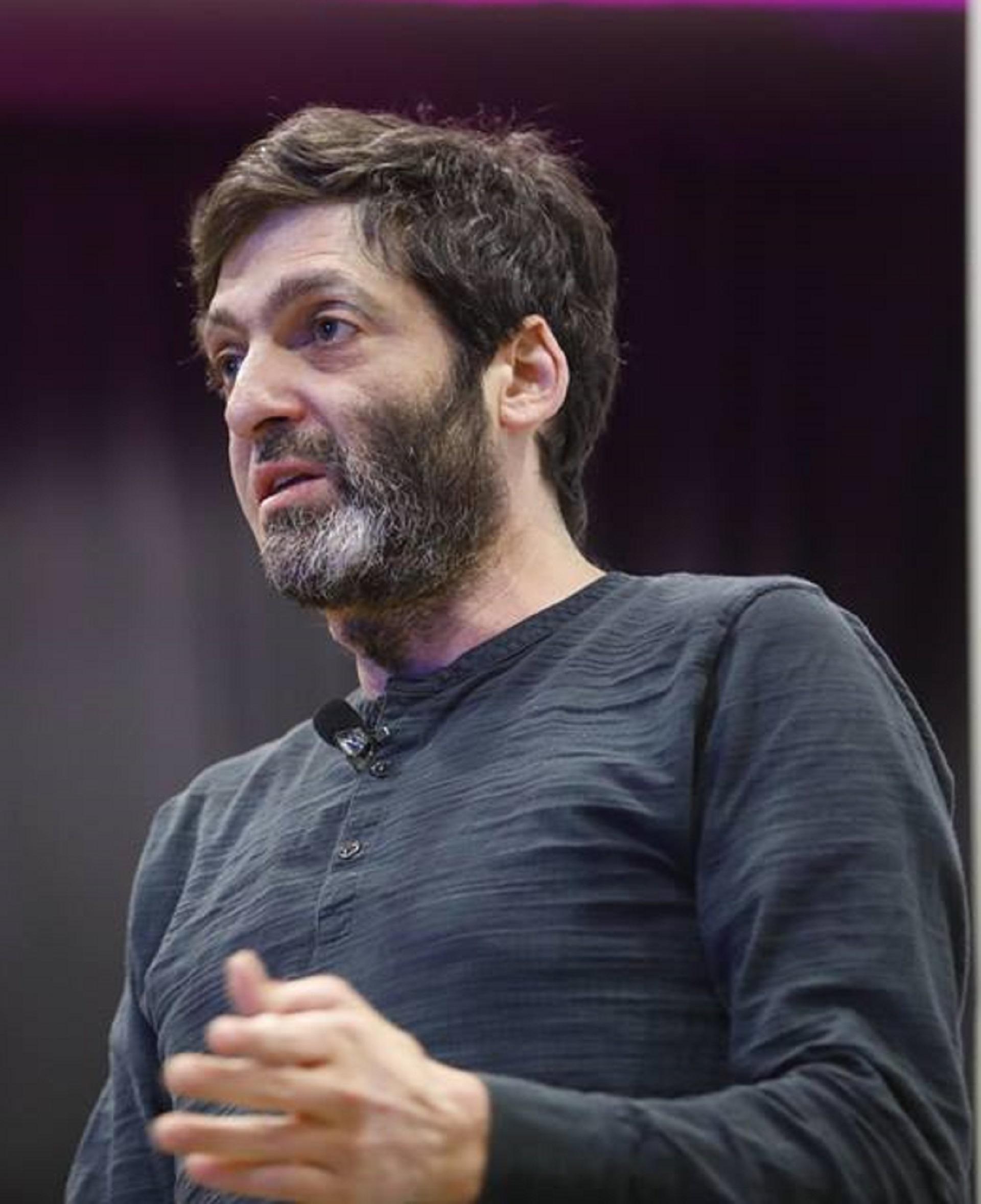 Dr Dan Ariely wears a dark blue long-sleeved shirt and holds his left arm at a 90 degree angle. He sports a beard with a patch of grey and looks intently at his audience.