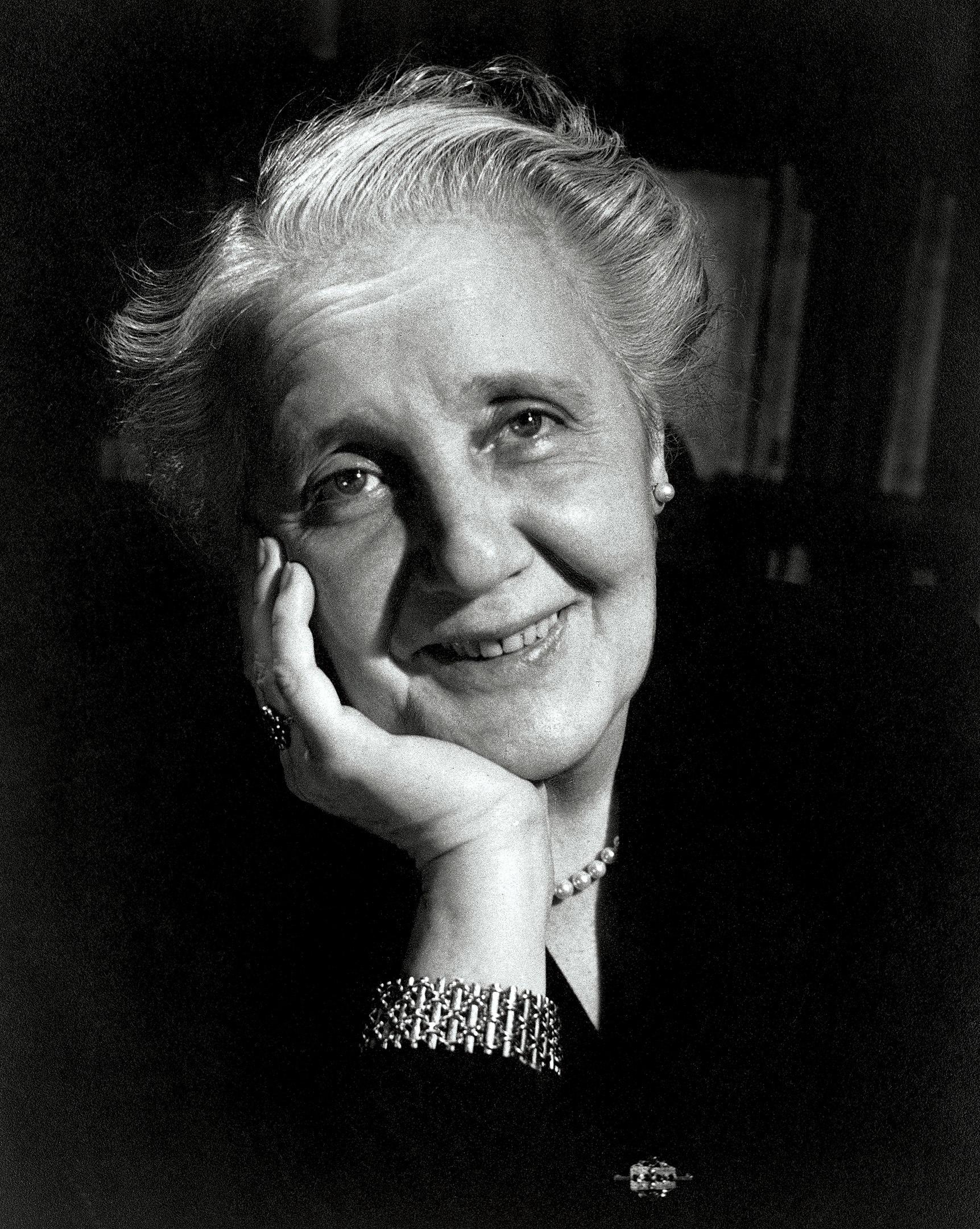 A greyscale image of Dr Melanie Klein with her silver hair swept back. Her right hand supports her chin and she smiles as she looks at the camera head-on.