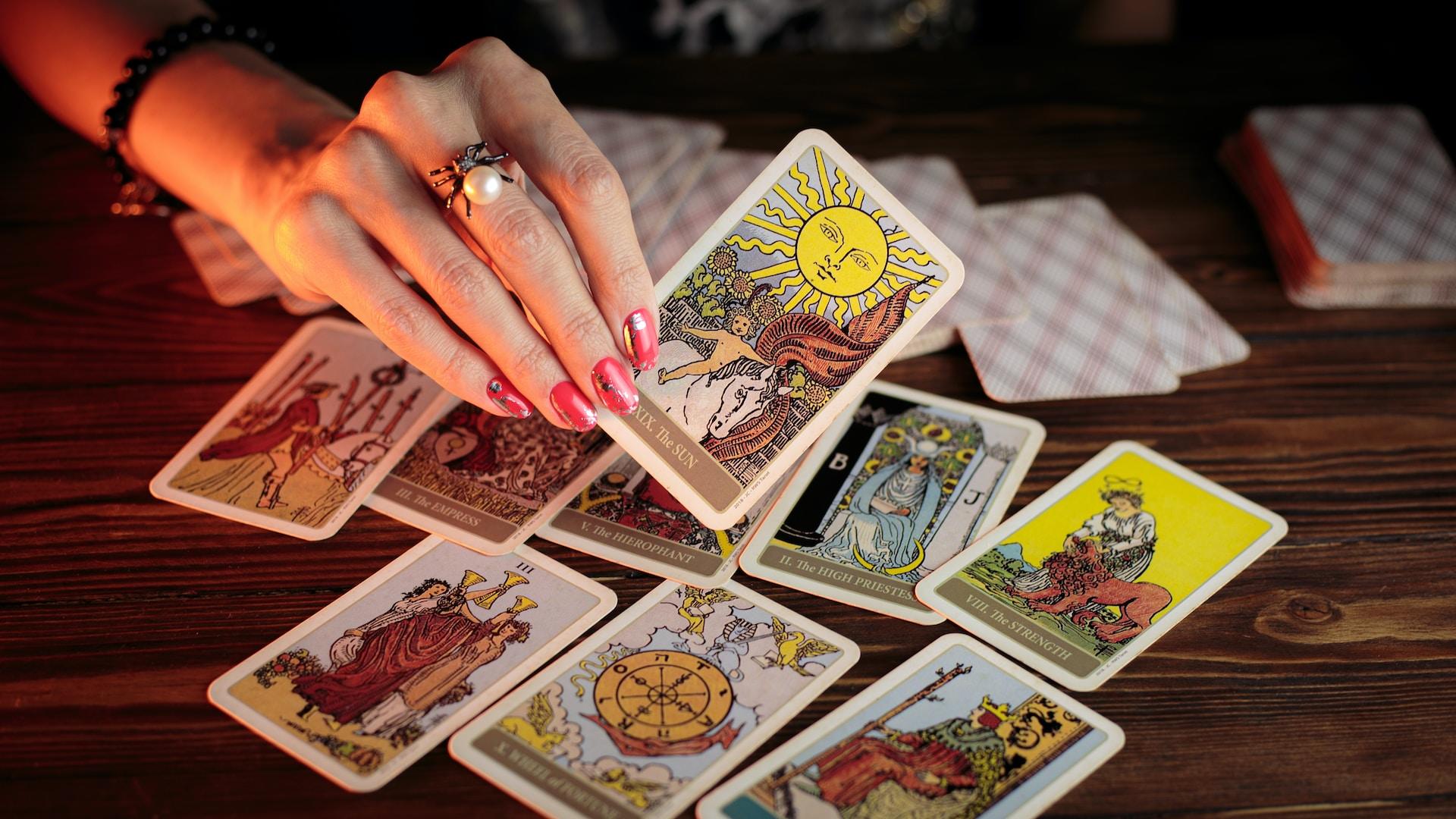 A hand with pink polish on its fingernails and a large pearl ring draws a tarot card from the pile scattered on a wooden table.