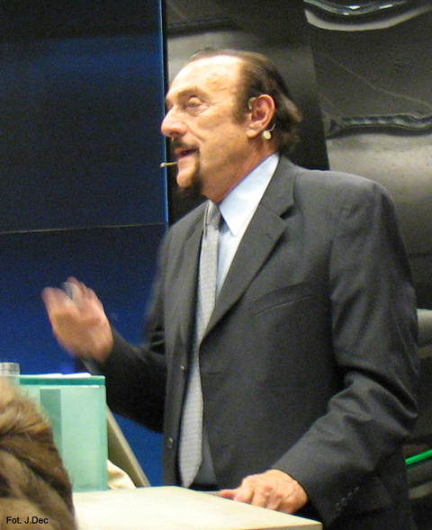 Dr Phil Zimbardo in 2009, dressed in a grey suit with a silver tie and white shirt. He wears a headset with a boom microphone and holds his right arm at a 90 degree angle, waving it to give emphasis to his speech.