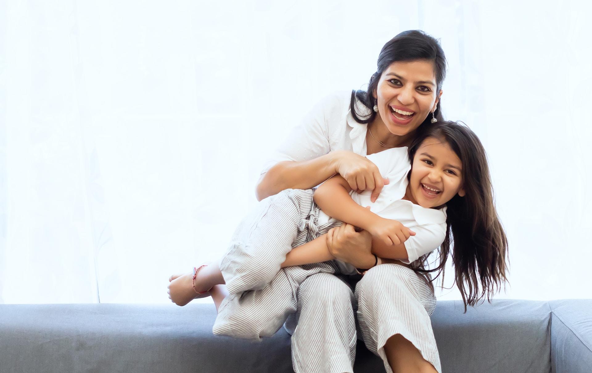 An adult wearing light-coloured clothing sits on a low wall holding a young, smiling child who also wears light-coloured clothing on their lap.