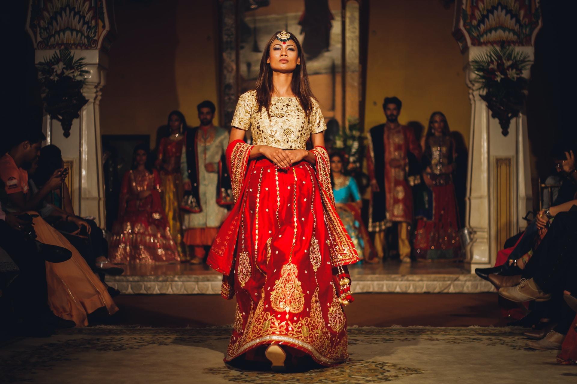 A woman in a red and gold costume stands in a great hall in front of a crowd of people, preparing to dance.