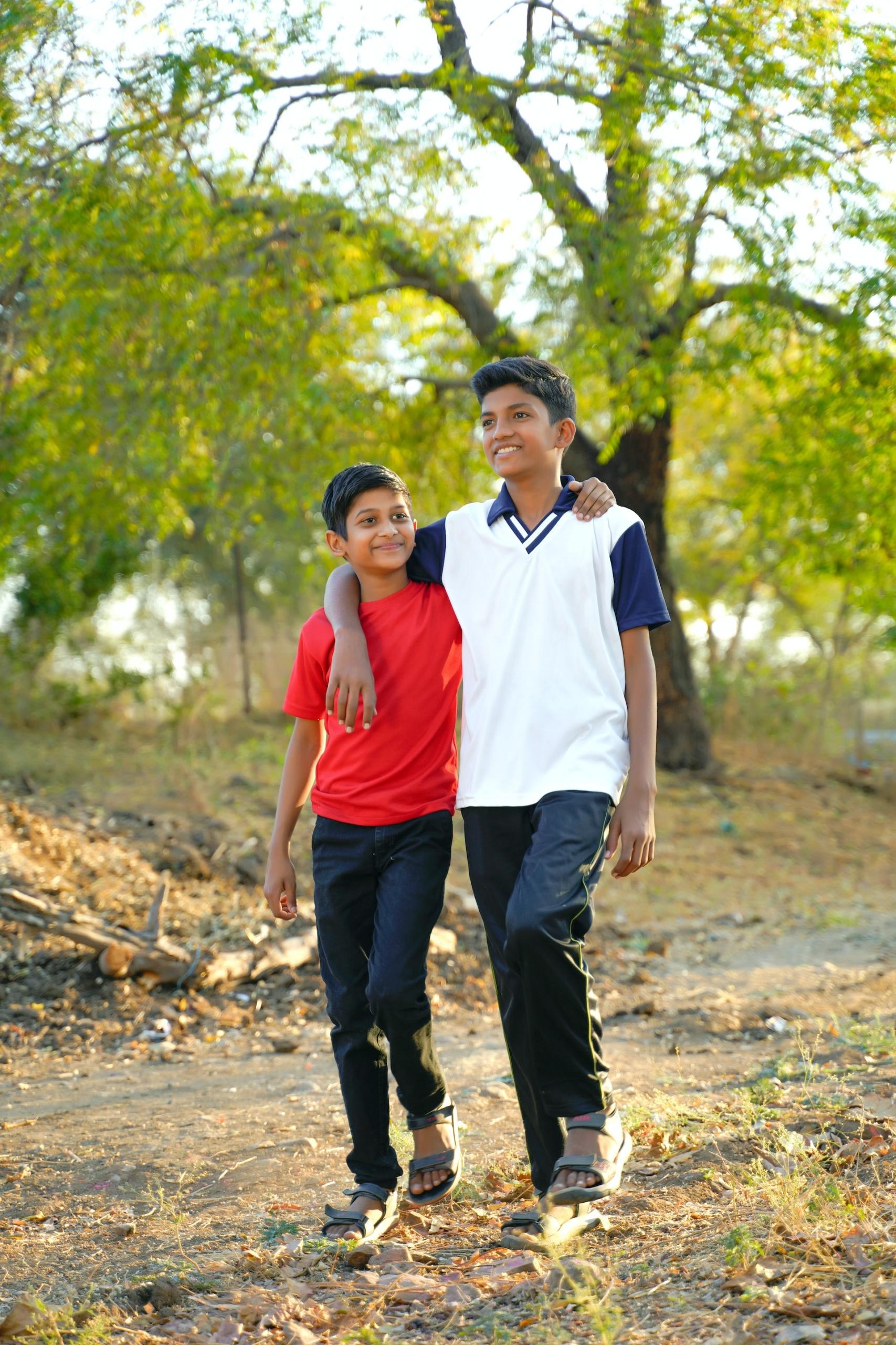 A boy wearing a white sleeveless knit vest with blue trim over a dark blue shirt and dark trousers walks arm in arm with a smaller boy who wears a bright red shirt and dark trousers.