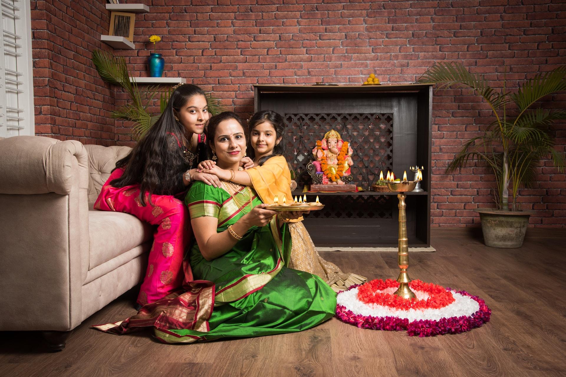 A woman in a green and gold sari sits on the hardwood floor holding a platter of small candles while her daughters hug her from either side.