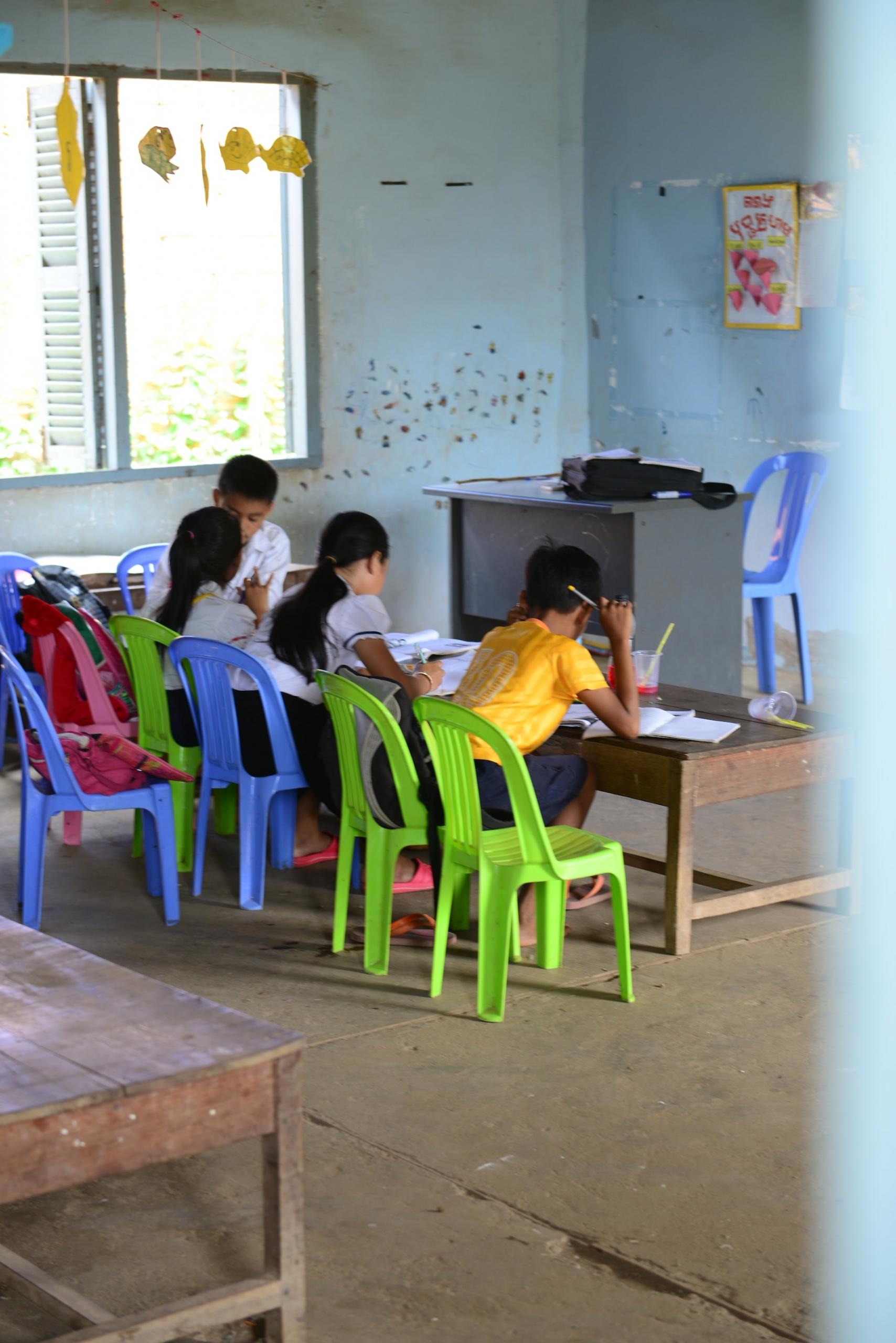 Three young students sit in colourful plastic chairs, bent over a low table as they do schoolwork. At the end of the row of tables, the teacher sits, talking with the last student in the row.