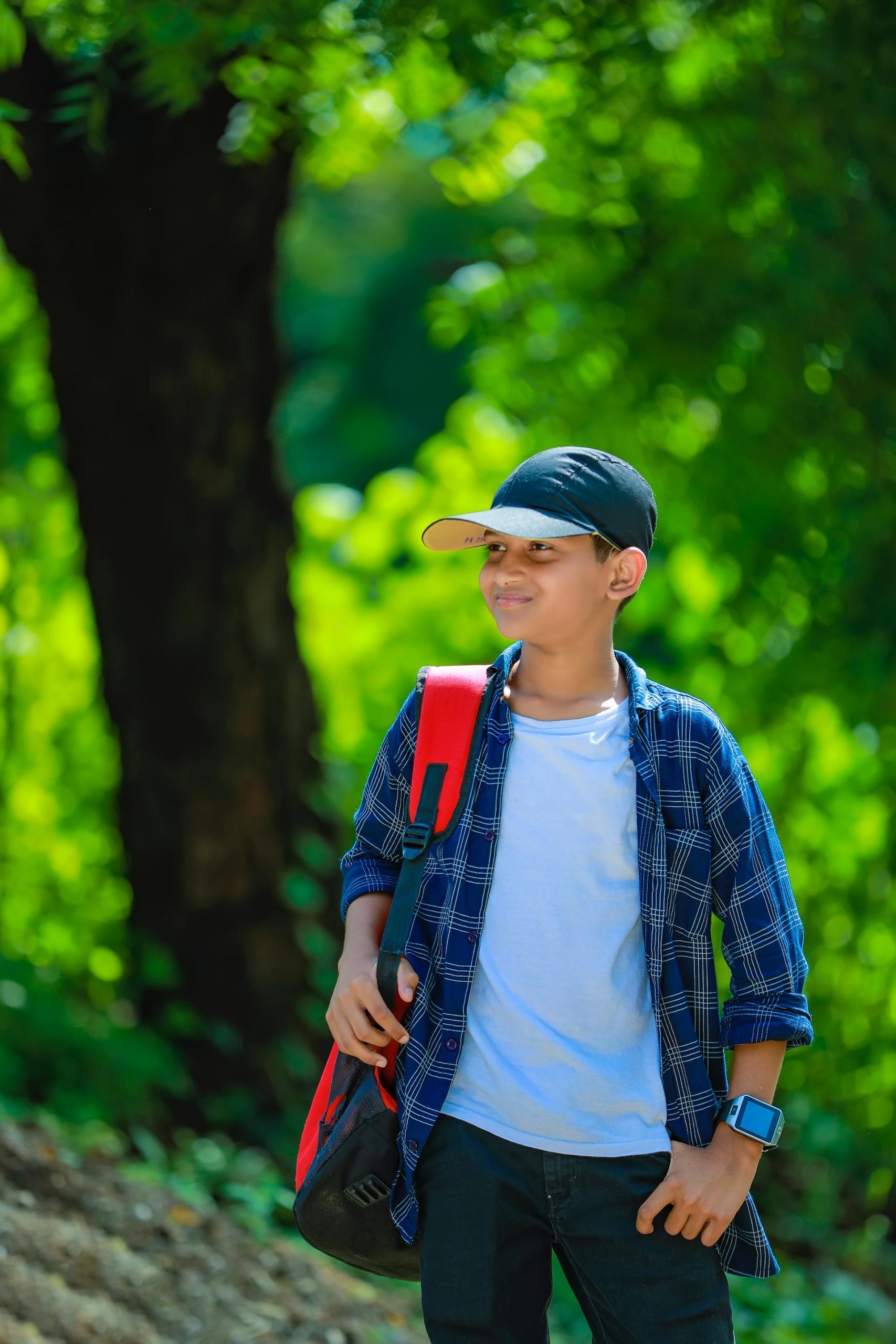 A boy wearing a blue ballcap, opened blue checked shirt over a white tee-shirt and dark trousers has a red and black backpack slung over his shoulder as he walks through a green area, smiling.