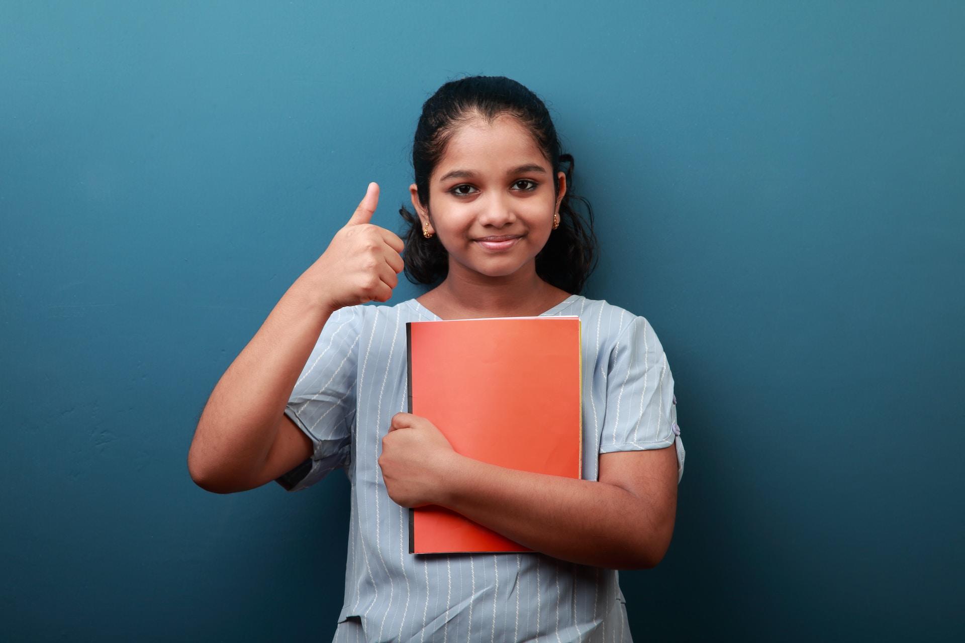 A student clutching an orange notebook to their chest wears a light blue, short sleeved top and makes a thumbs-up sign as they stand in front of a chalkboard and smiles.
