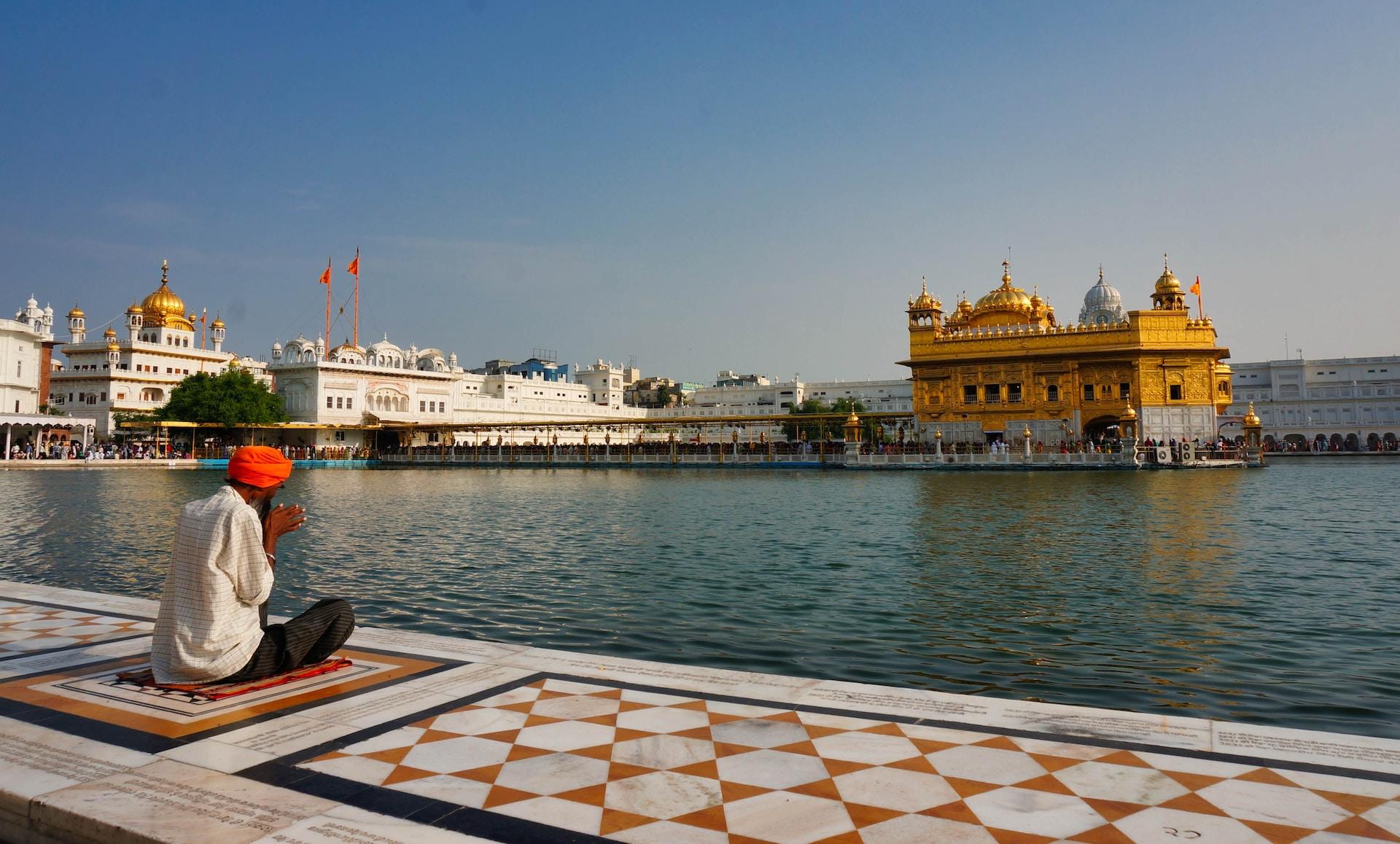 A sikh temple with a golden facade across the water, with a devout wearing a red turban and light coloured clothing praying on the opposite bank.