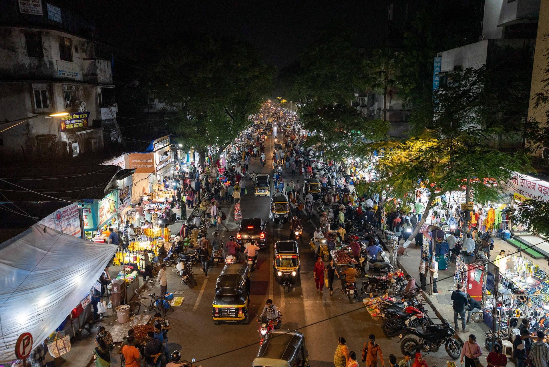 A busy Mumbai street at night, with tuk-tuks, cars, buses and pedestrians all crowding the street and the shops brightly lit.