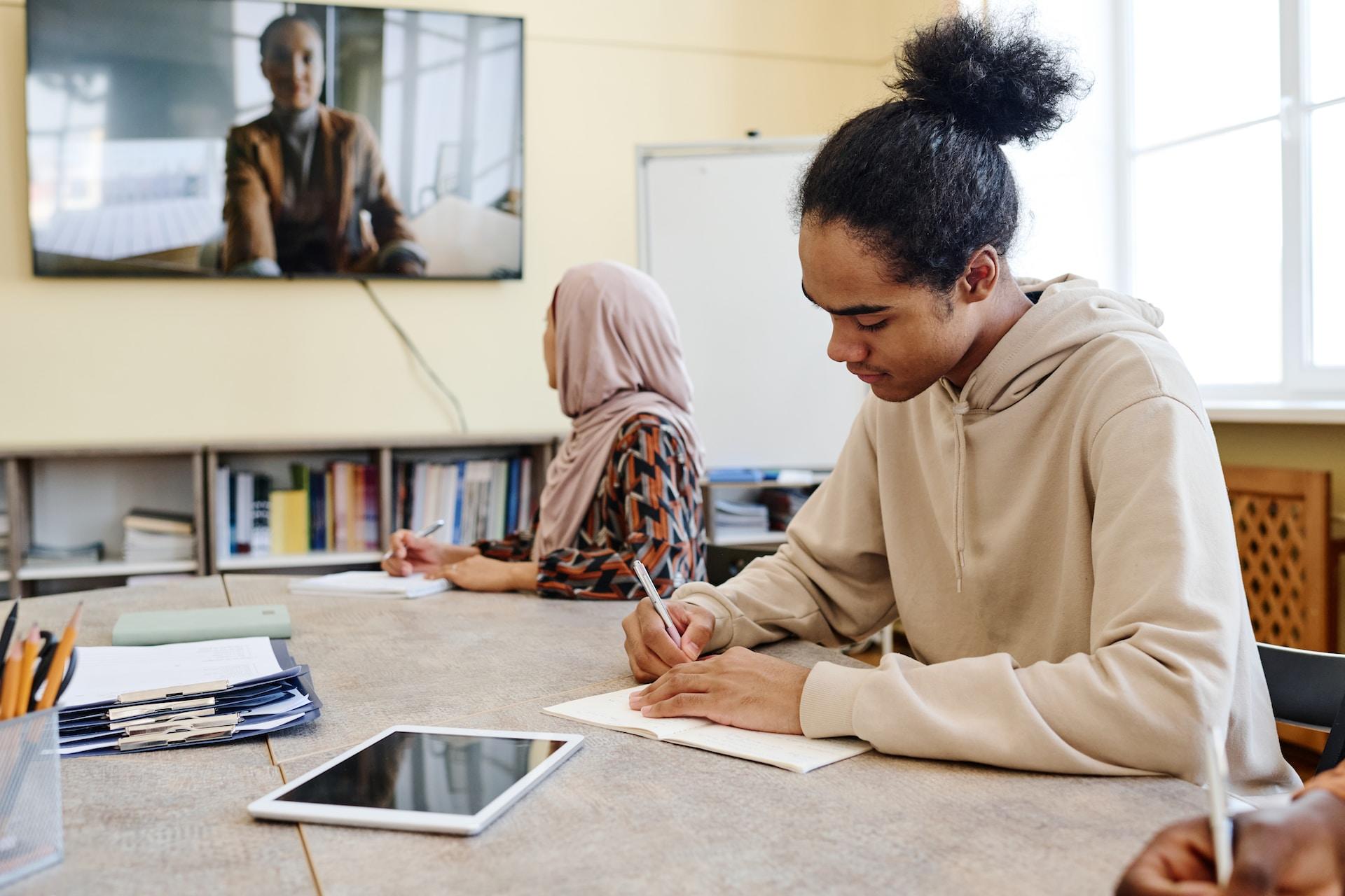 A person wearing a light coloured top sits at a table and writes while a woman wearing a light-coloured hijab watches the teacher on the wall mounted monitor.