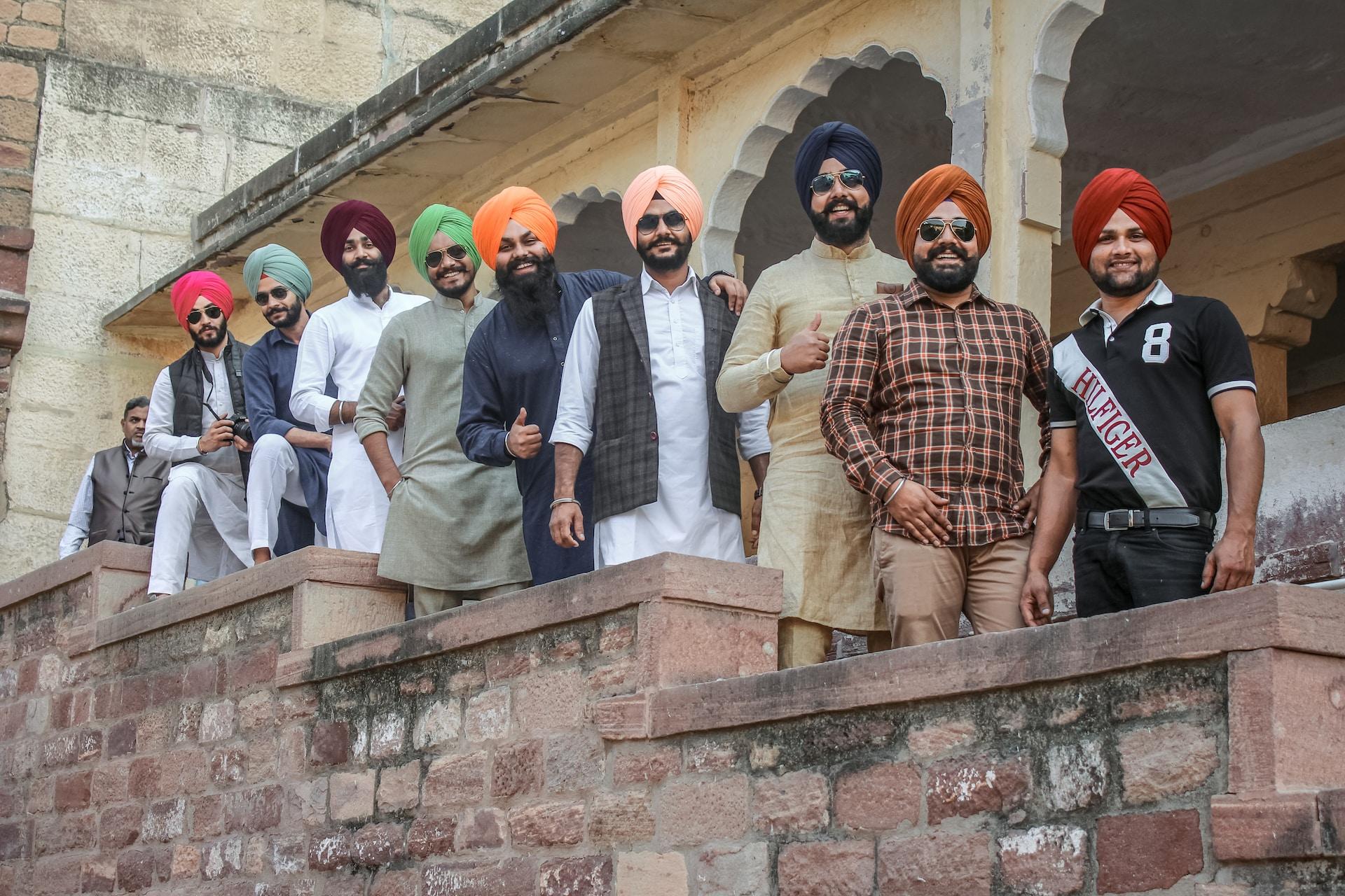 Nine Sikh men posing along a low parapet wall that borders a staircase outside of a stone building.