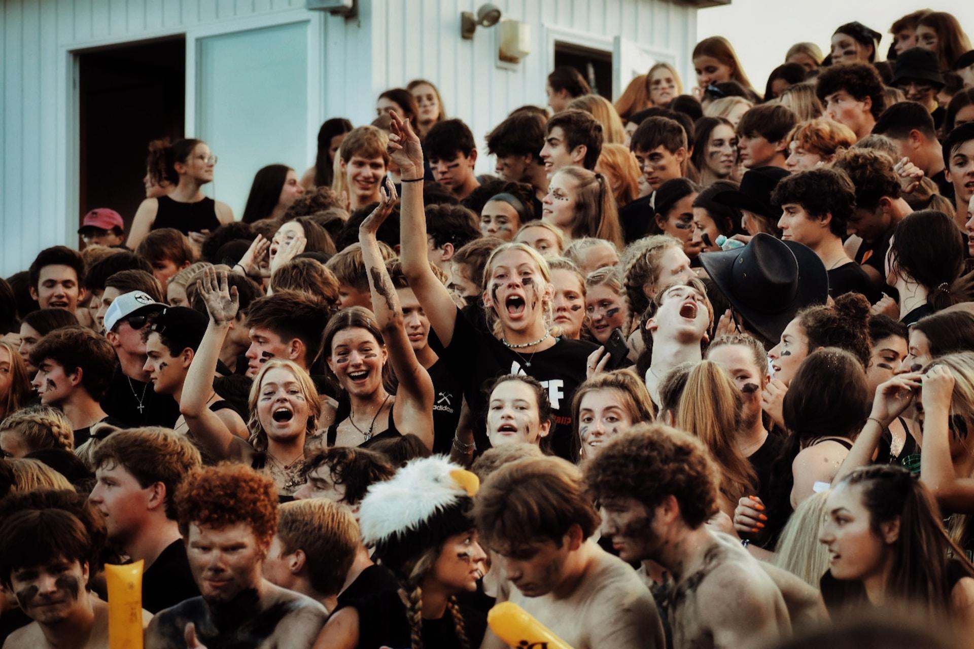 A student body clustered tightly together with some wearing face paint and others headdresses, cheering as though they were at a sports rally indoors.