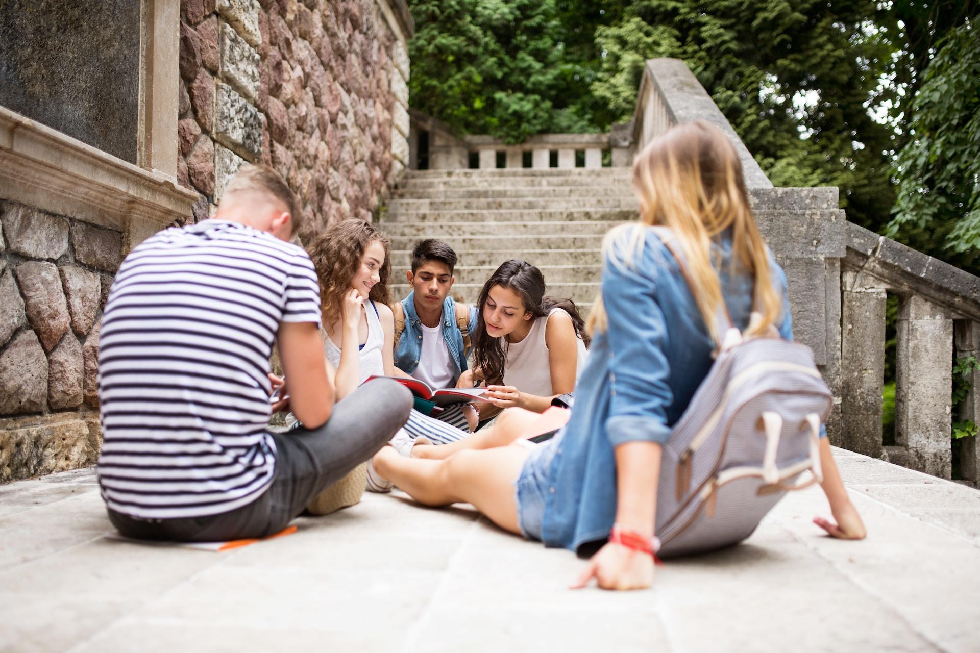 Four young people sit outdoors on a stone walkway next to a sandstone wall , in front of a staircase on a sunny day.