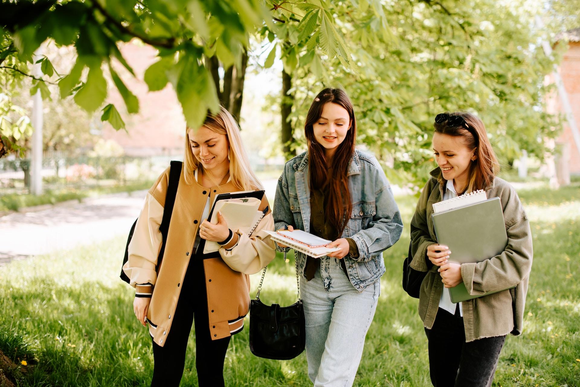Three women walk together in a greenery area, wearing Autumn clothing and carrying school books and bags. 