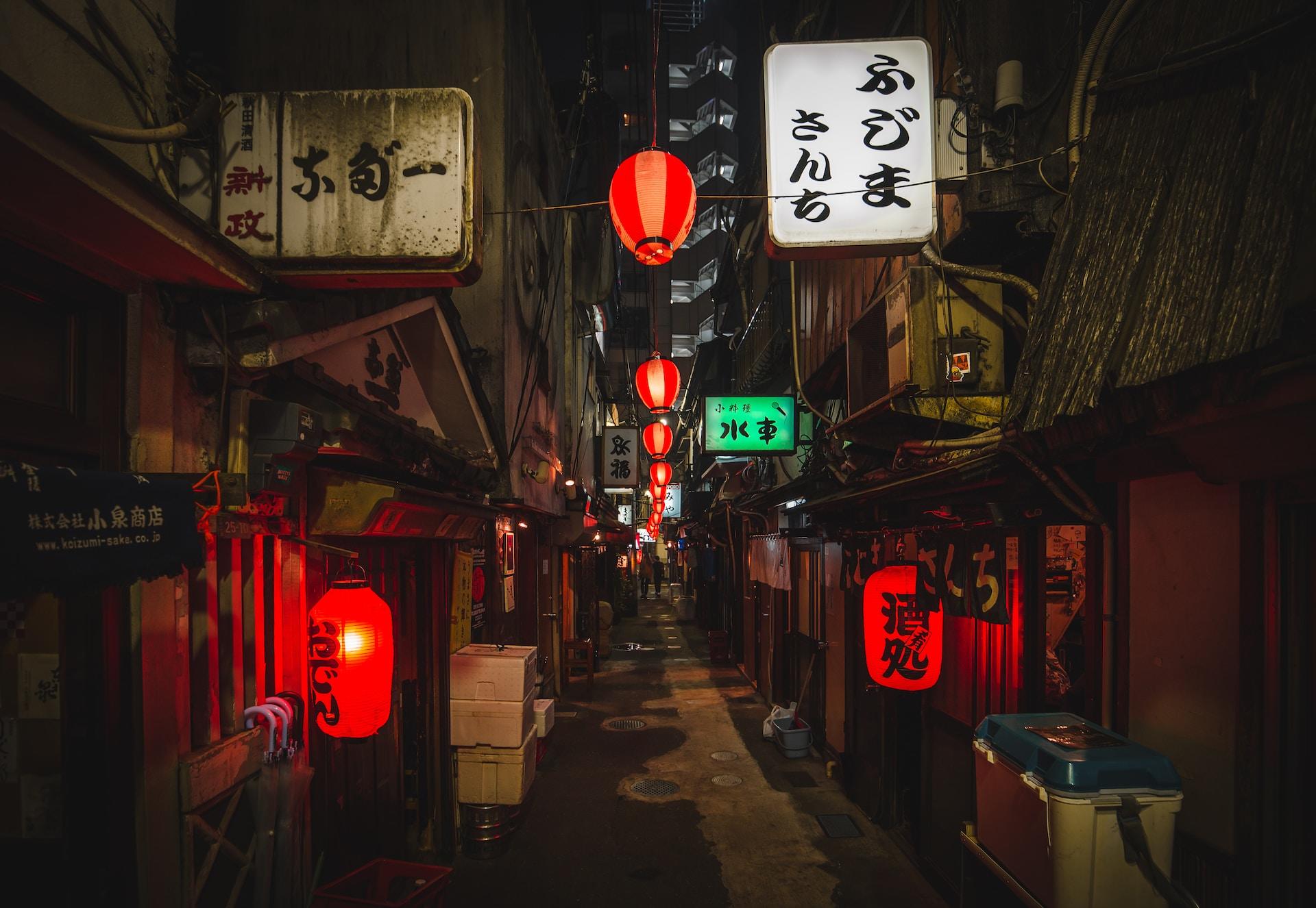 A narrow alley in Japan's oldest district at night, illuminated with red lanterns featuring writing in black and other signs in various colours. 