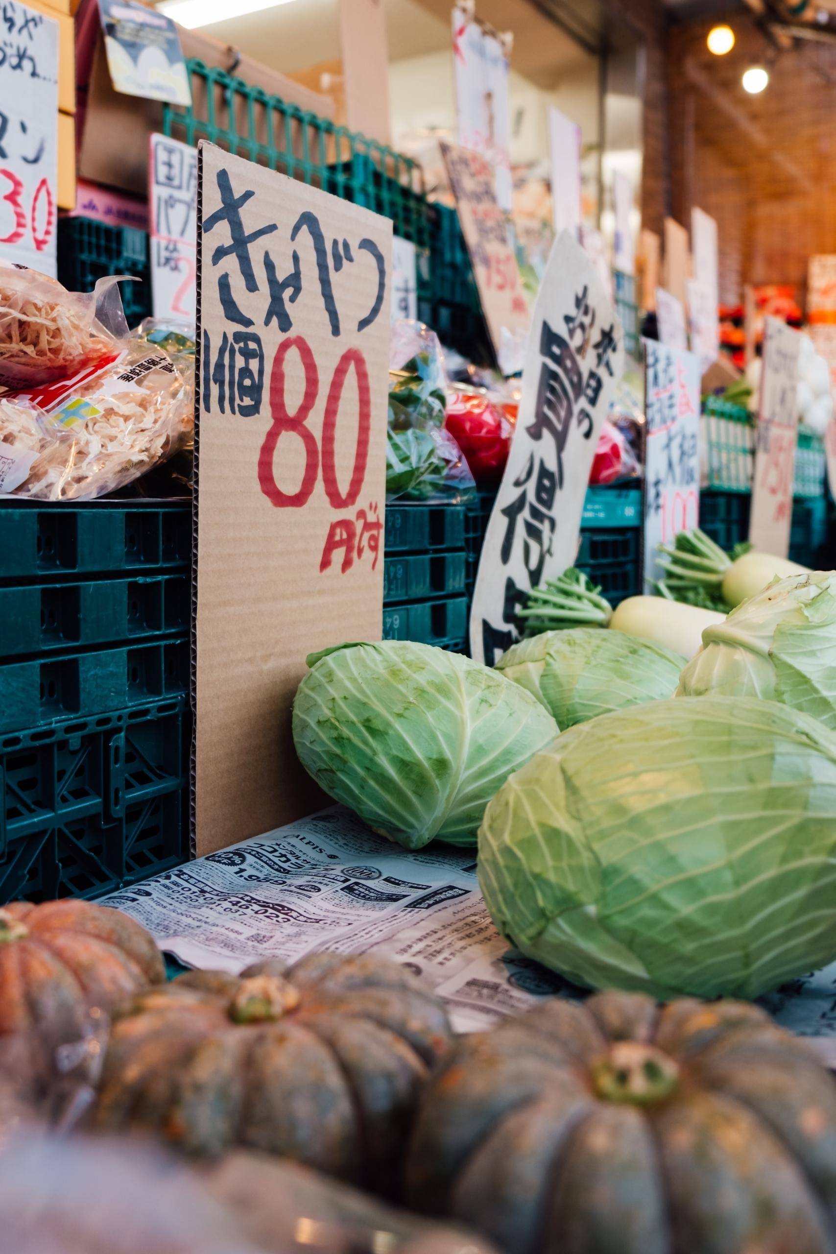 A Japanese farmer's market with cabbages and gourds on display and a cardboard sign with the produce's name written in black and their prices written in red ink. 