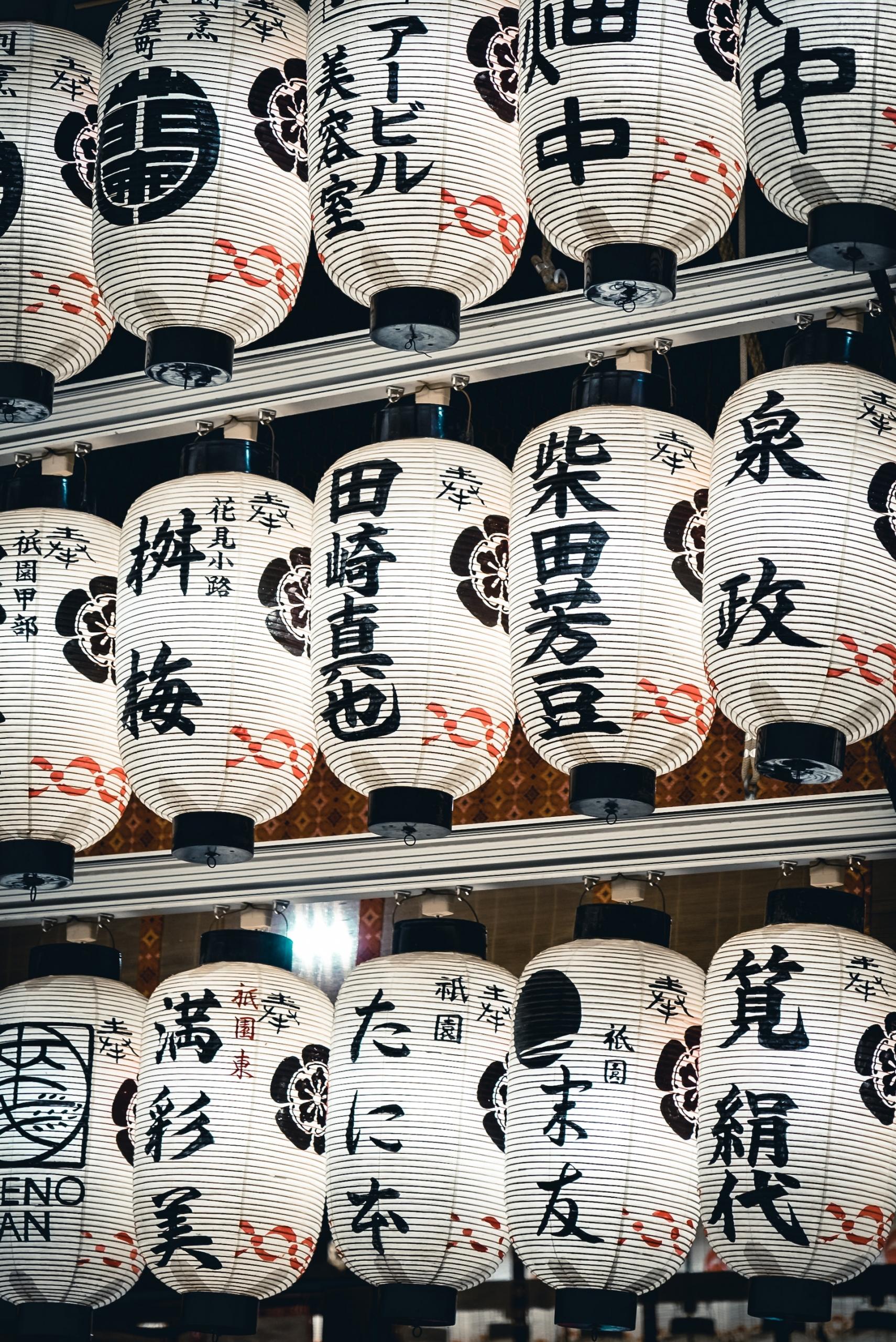 Three rows of white paper lanterns with Japanese pictograms painted on them in black ink, and a red frieze at the bottom of each lantern. 