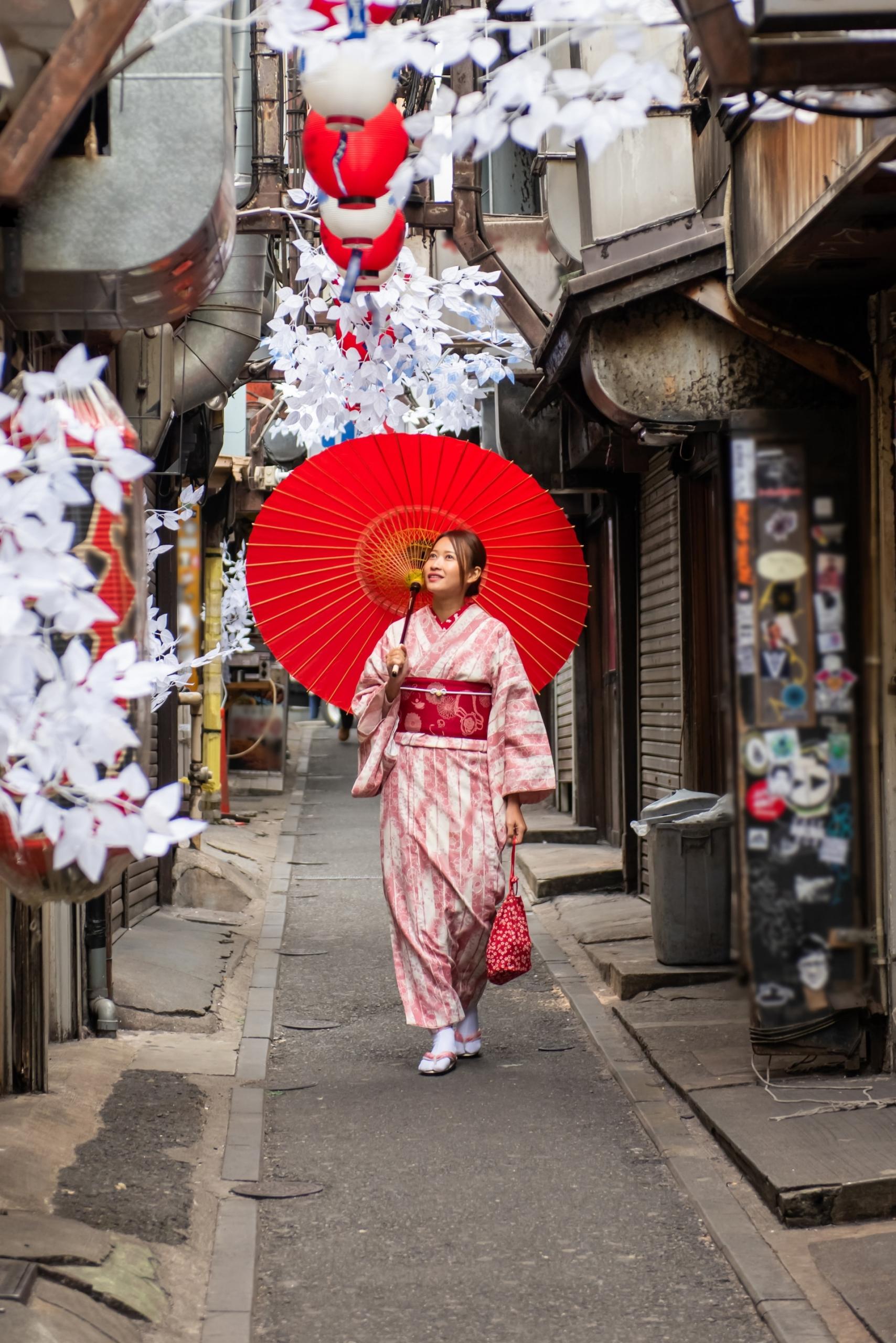 A young woman wearing a pink kimono with a red obi and carrying a red parasol walks down a narrow alleyway overhung with white and red decorations. 